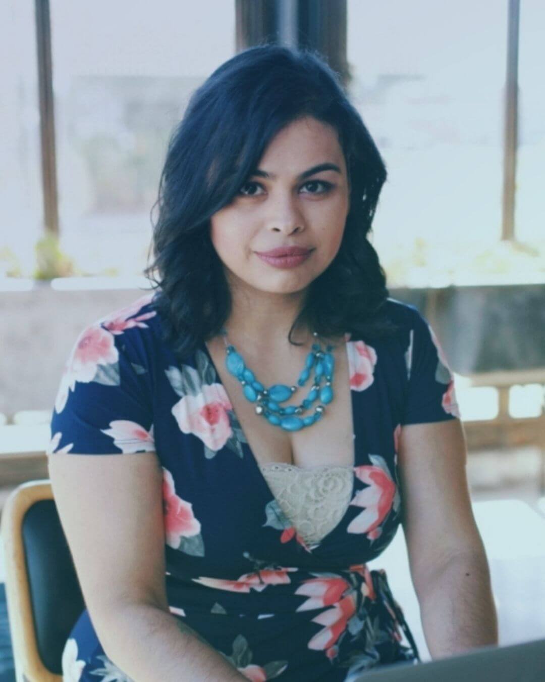 A woman with shoulder-length black hair, wearing a navy blue dress with pink and white floral patterns, and a blue beaded necklace, sitting indoors near a window.