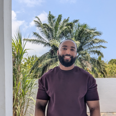 A smiling man with a beard standing outdoors in front of palm trees and greenery under a partly cloudy sky.