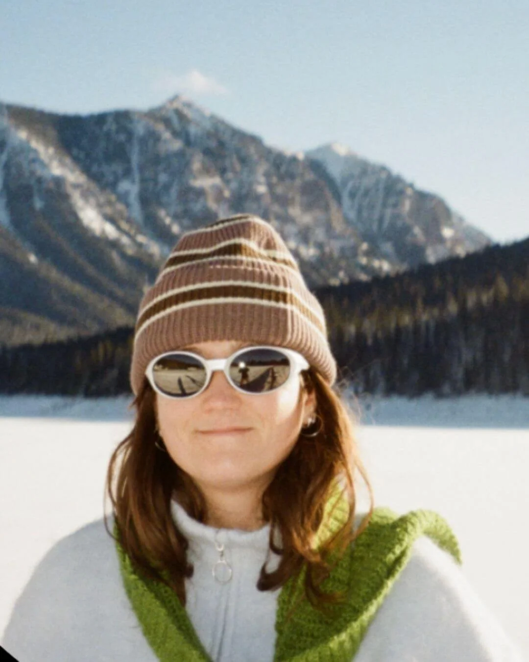 A person wearing sunglasses, a striped beanie, a white zip-up sweater, and a green vest standing outdoors in a snowy landscape with mountains in the background.