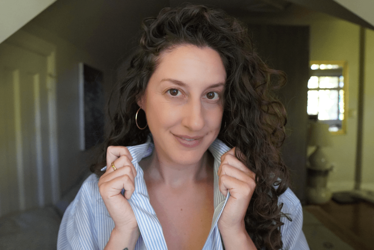 A woman with dark, curly hair wearing a blue and white striped shirt and hoop earrings, smiling and holding her collar.