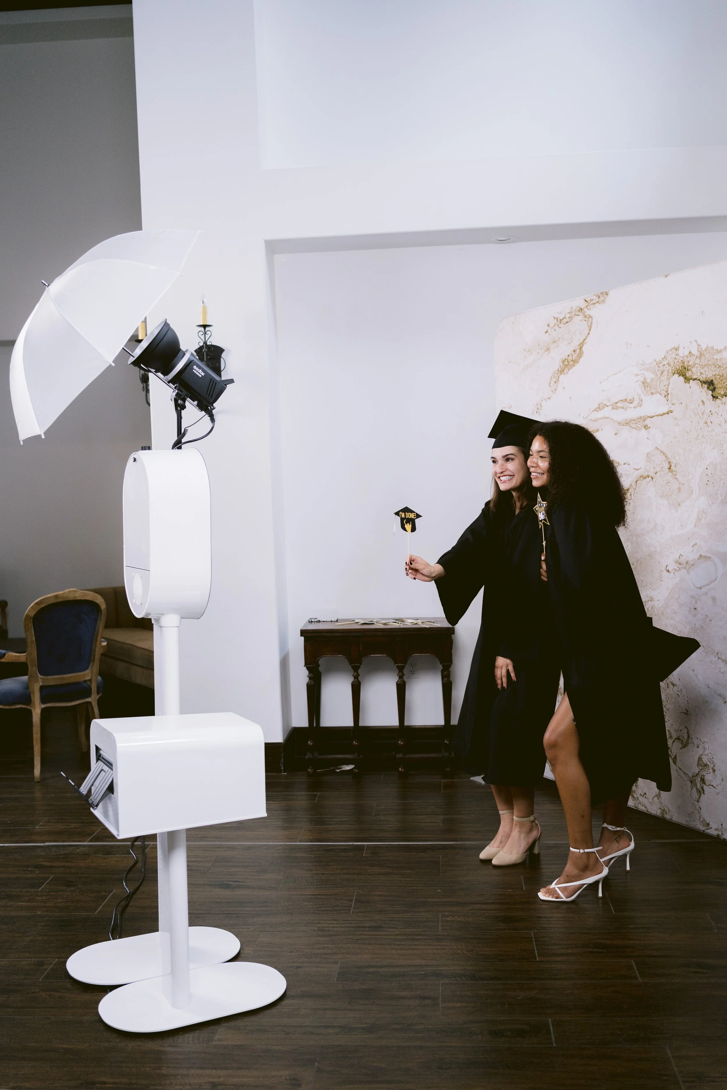 Two women in black graduation gowns and caps celebrate at a graduation photo shoot, holding up props with congratulatory messages, in a room with a marble accent wall and dark wood flooring.
