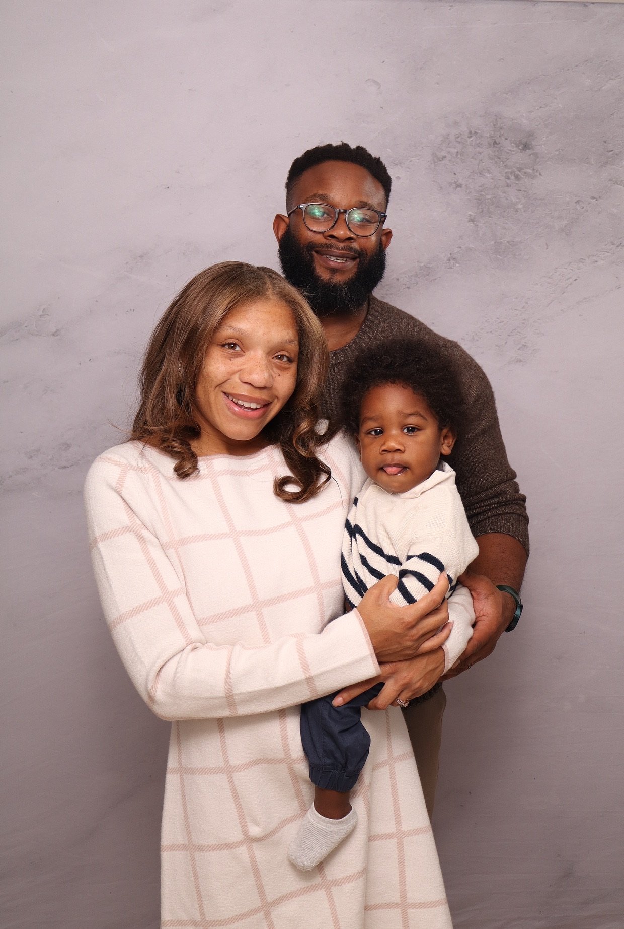A family of three, consisting of a woman, a man, and a young boy, standing together in front of a plain gray wall, smiling and posing for the photo.