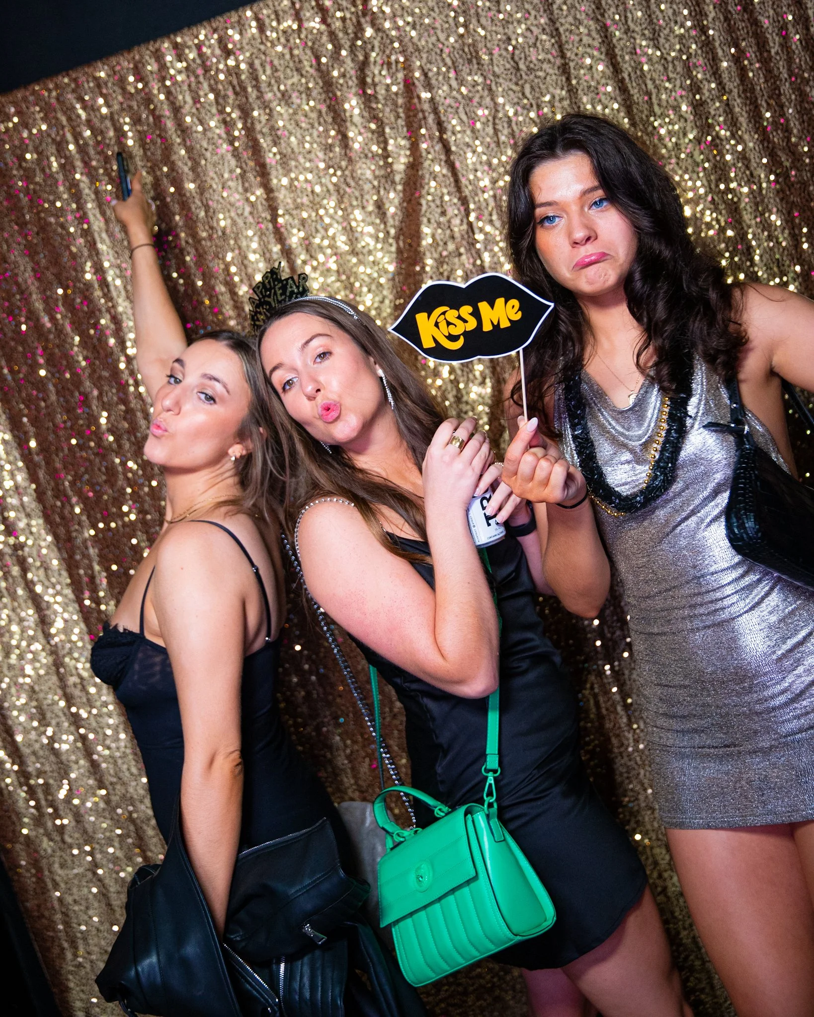 Three women posing in front of a gold sequin backdrop at a party or event. They are making playful facial expressions and holding props, including a speech bubble sign that says 'Kiss Me'. The women are dressed in stylish outfits with accessories lik