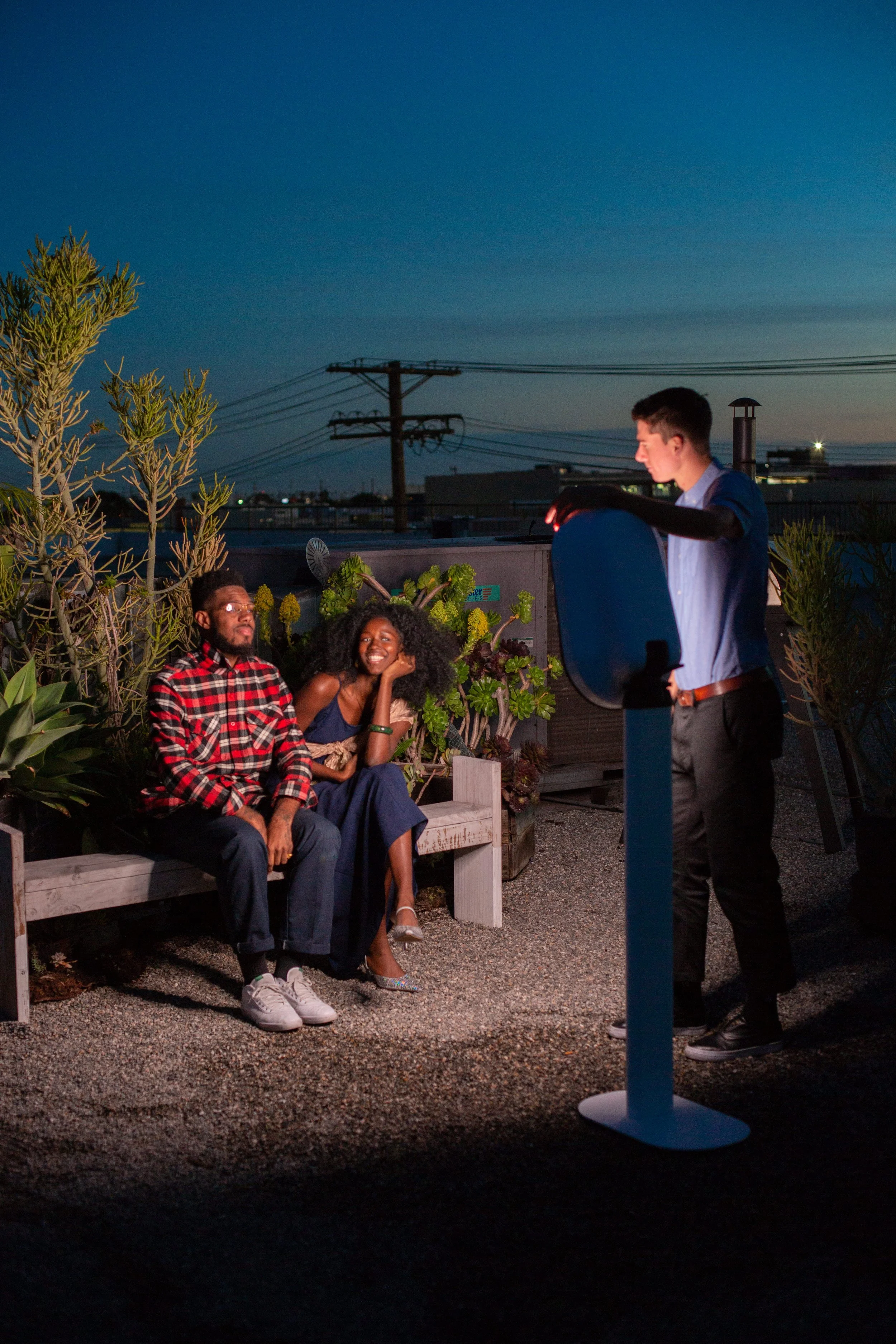 Three people on a rooftop; two sitting on a bench, in front of CLTPhotoBoothMagik’s Selfie Booth, surrounded by plants and urban scenery.