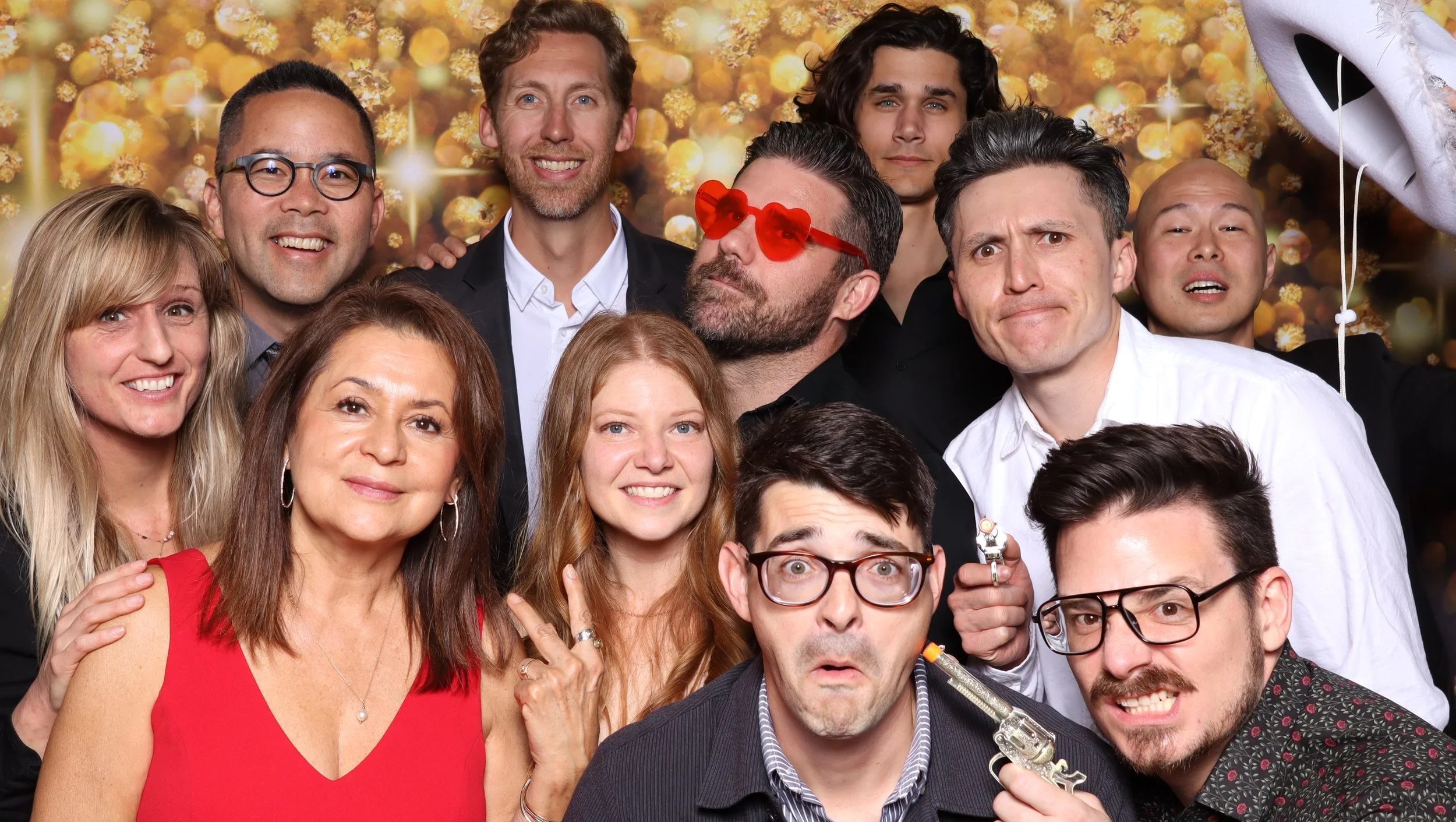 Group of smiling people in a photo booth with a gold glitter background, some wearing fun accessories like heart-shaped sunglasses and holding props.