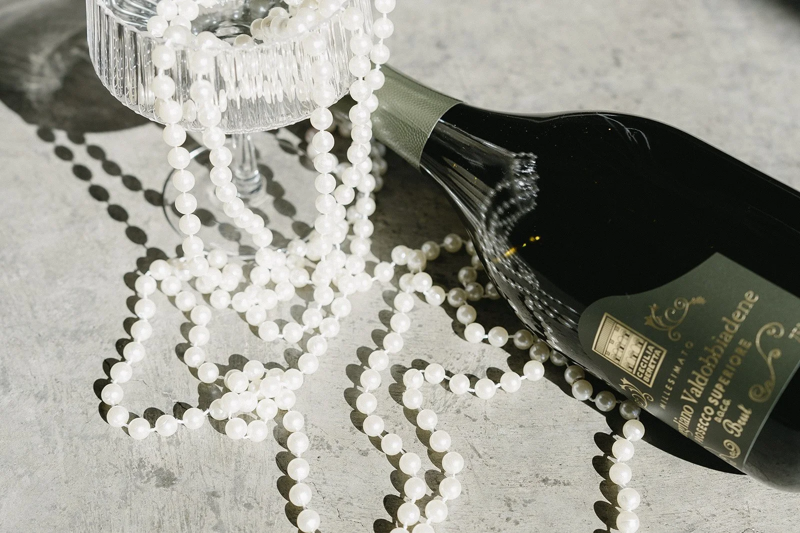 A wine bottle and a string of pearls draped over a clear glass cup on a concrete surface.