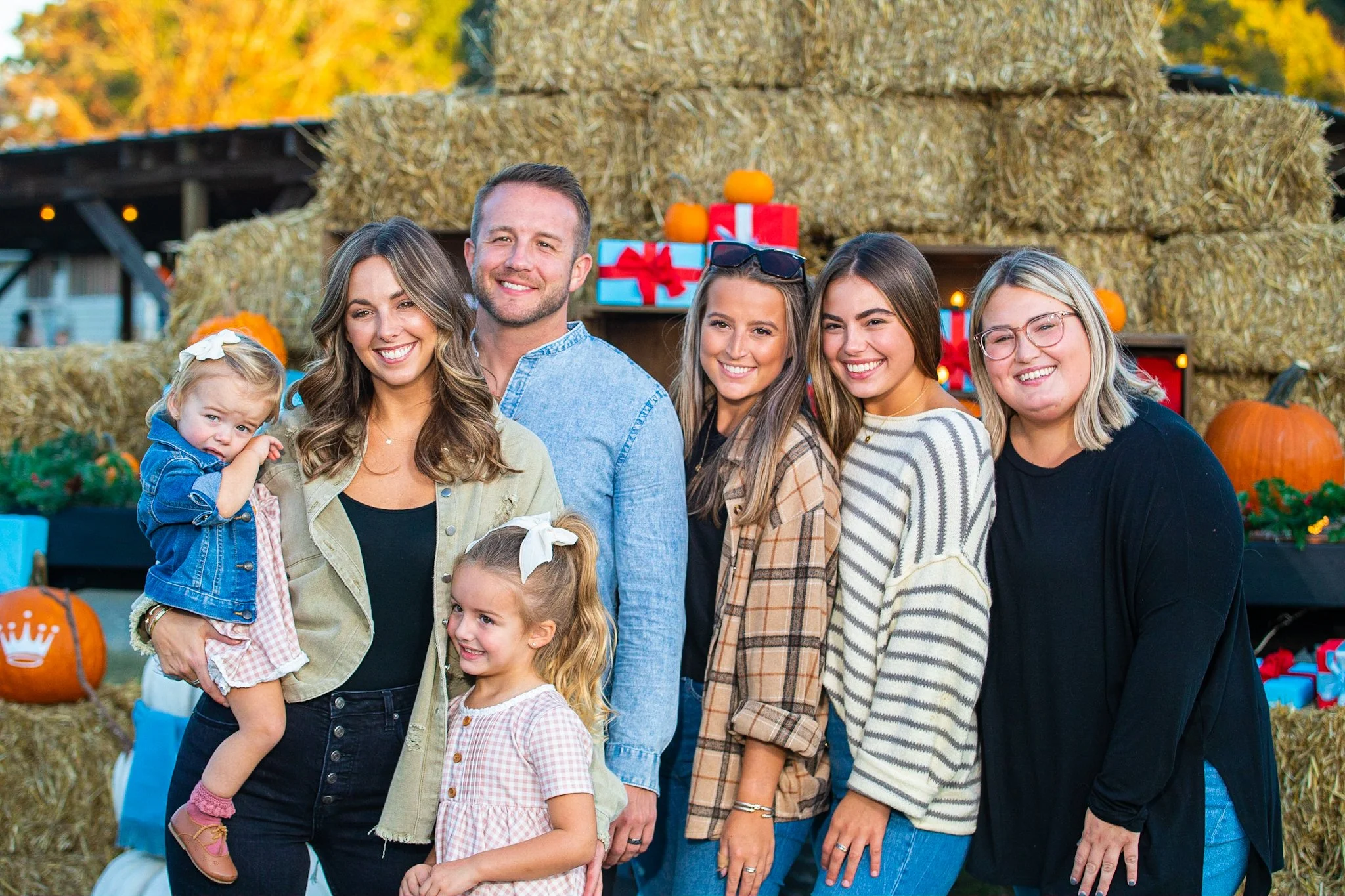 A family group of six people, including two young children and four adults, standing outdoors with hay bales, pumpkins, and gifts in the background, during fall.