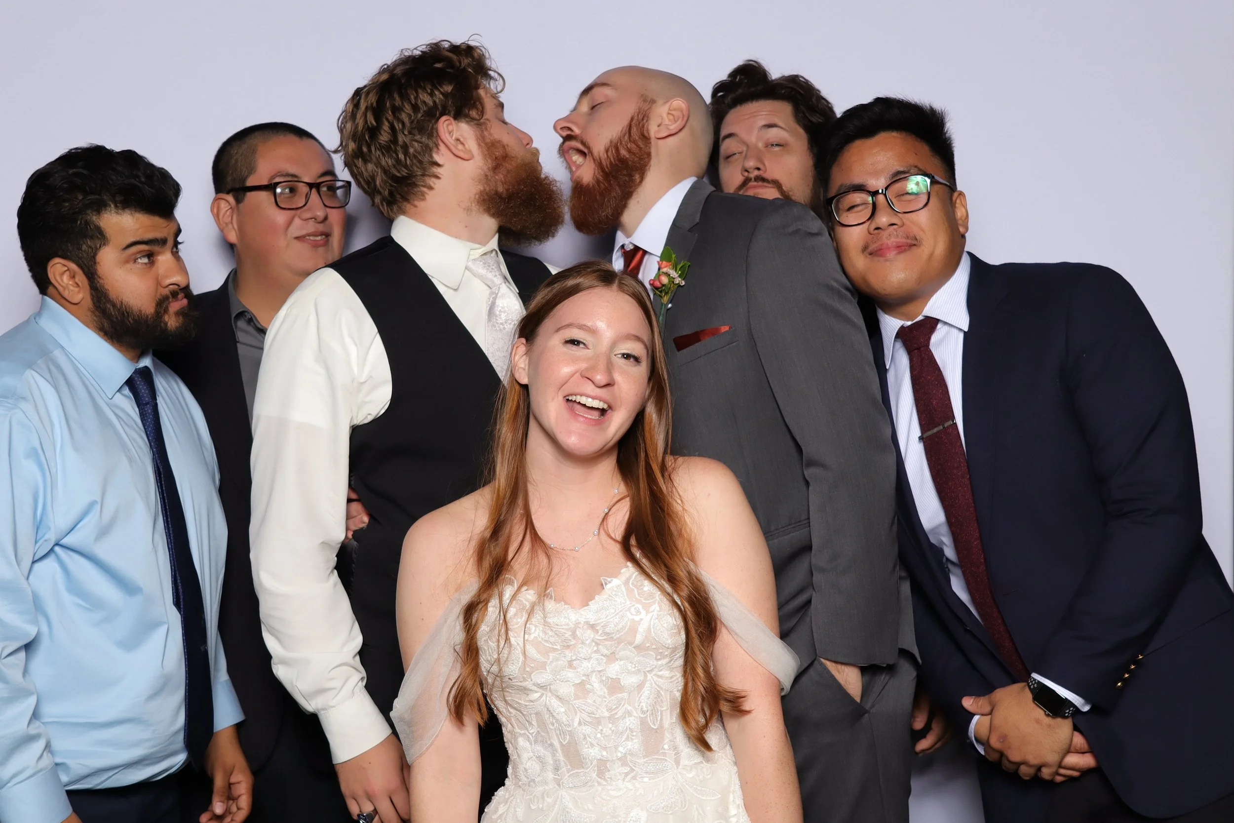 Group of diverse people in suits and dresses posing at a wedding celebration, with two men leaning in for a kiss.