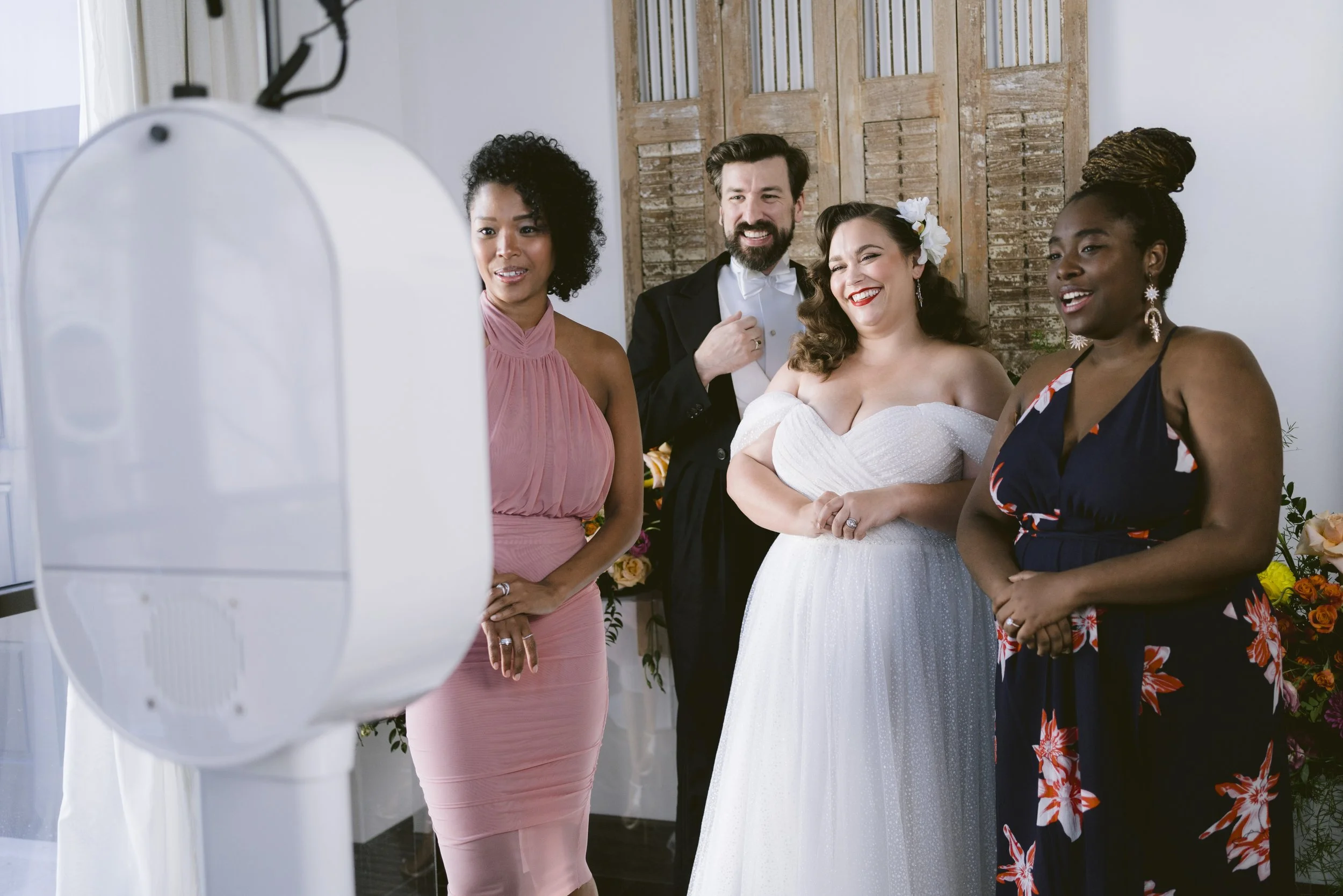 People gathered around a mirror during a wedding preparation, including a bride in a white dress with flowers in her hair, and three women in colorful dresses, with a wooden door in the background.