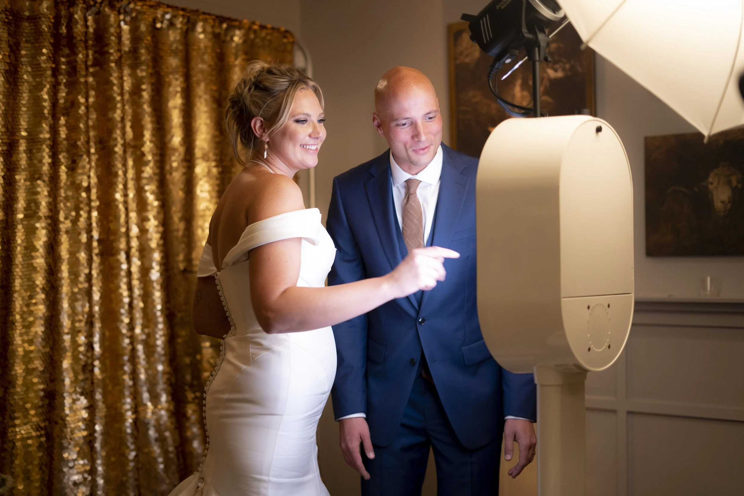 A woman in a wedding dress and a man in a suit smiling while looking at a photo booth at a celebration.