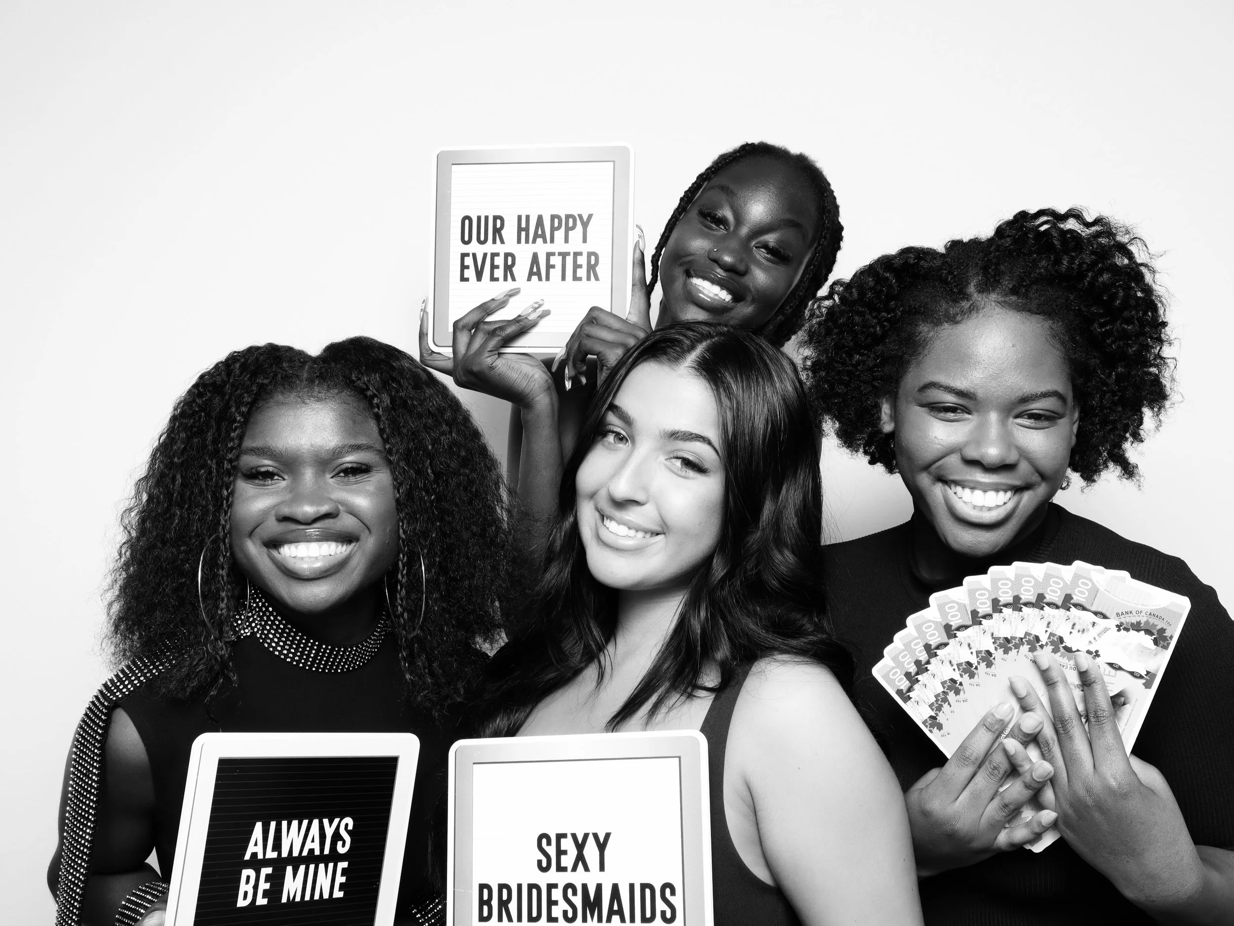 A group of diverse women smiling, holding signs that read "ALWAYS BE MINE," "SEXY," and "SEXY BRIDESMAIDS." One woman is holding a fan of Canadian hundred-dollar bills, and another is holding a tablet. One woman is holding a sign that says "OUR HAPPY EVER AFTER."
