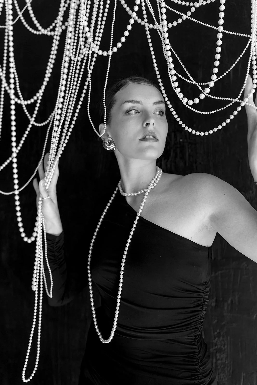 Woman in black dress with pearl necklaces and earrings, looking up.