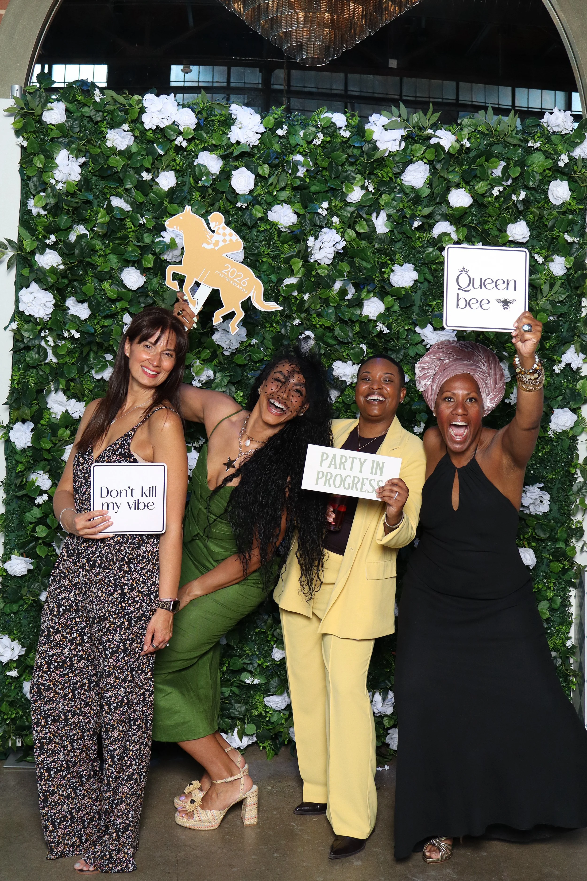 Four women smiling and posing in front of a greenery wall with white flowers, holding various signs. One woman on the right is wearing a large pink turban and a black dress, holding a sign that says 'Queen bee'. The second woman from the right is in a light yellow suit with curly hair, holding a sign that says 'Party in Progress'. The third woman from the right is wearing green, with long curly black hair and glasses, holding a sign that says 'Don't kill my vibe'. The woman on the far left is wearing a printed jumpsuit with floral pattern.