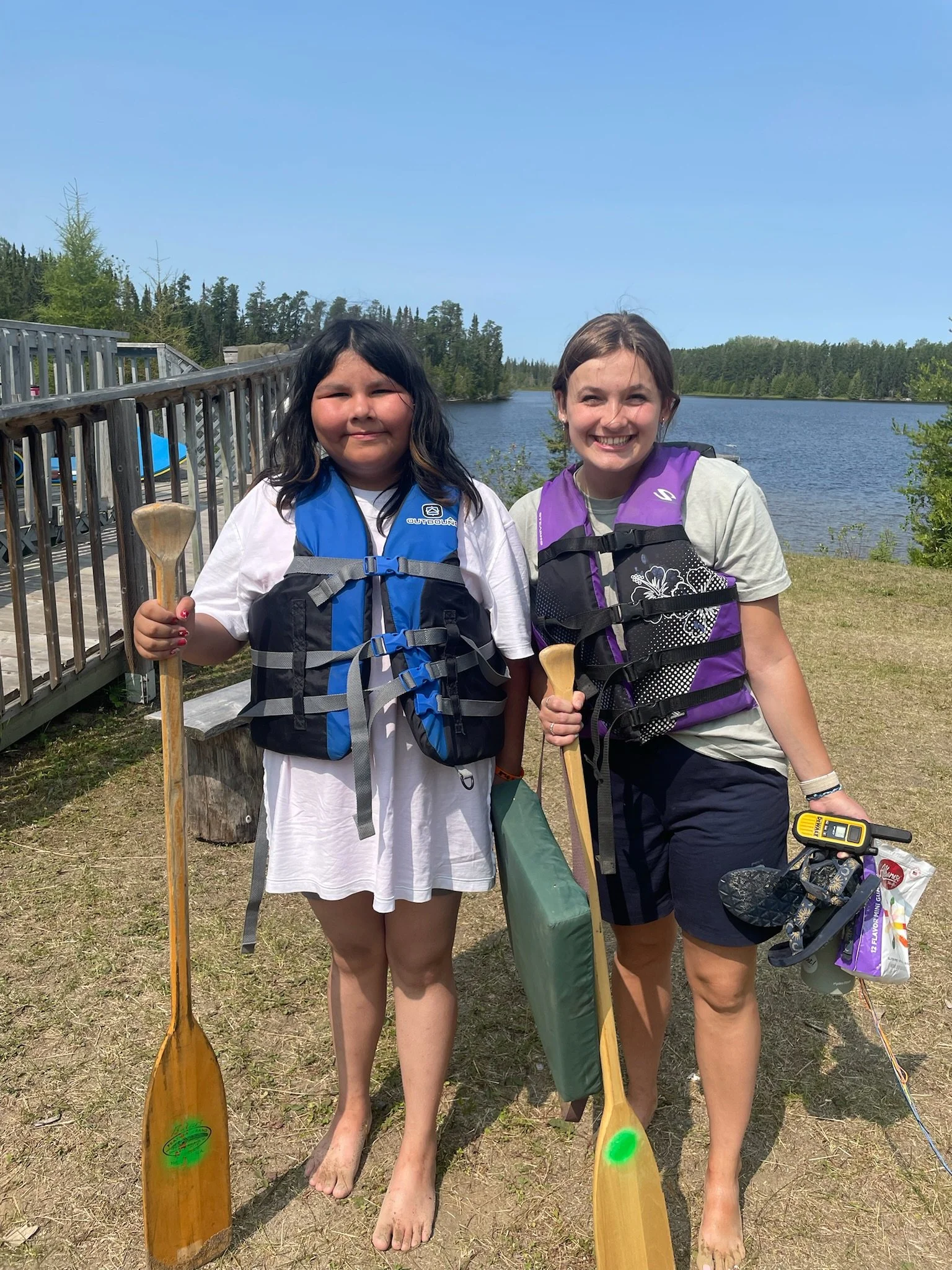 Two people wearing life jackets holding wooden paddles, standing by a lake with trees in the background.