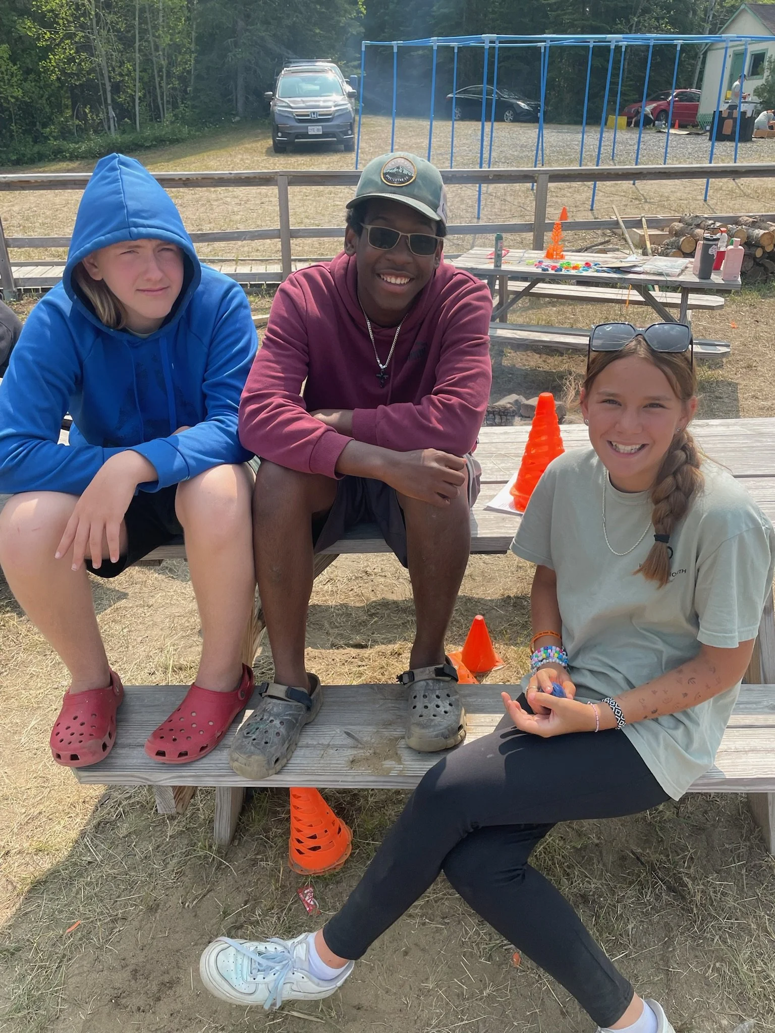 Three people sitting on bleachers, with a wooded area and parked cars in the background. Two are wearing casual clothing and Crocs, and one has a hood up. They appear to be outdoors, possibly in a recreational or camp setting with orange cones nearby.