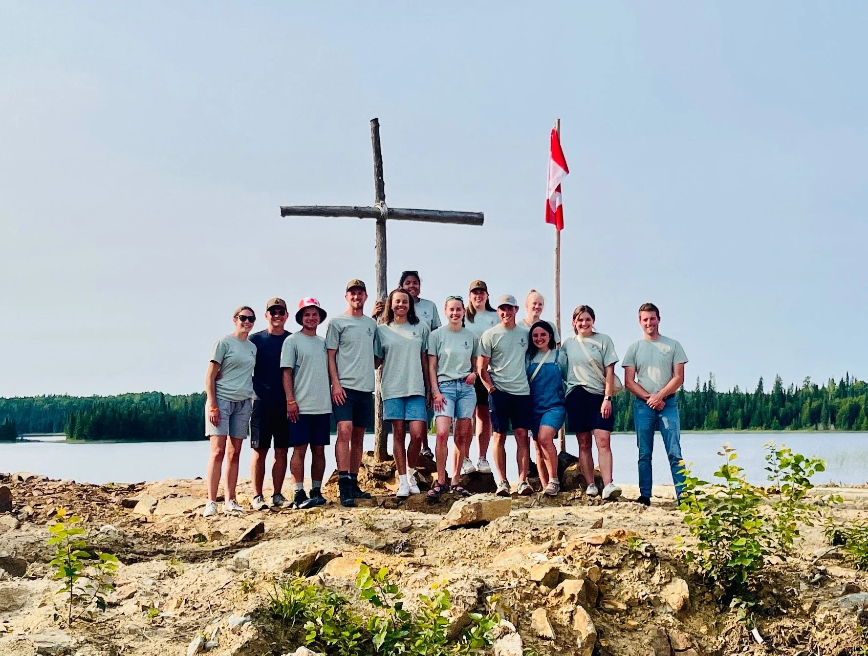 Group of people wearing similar shirts standing on rocky lakeside with a wooden cross and Canadian flag in the background, surrounded by trees and a lake.