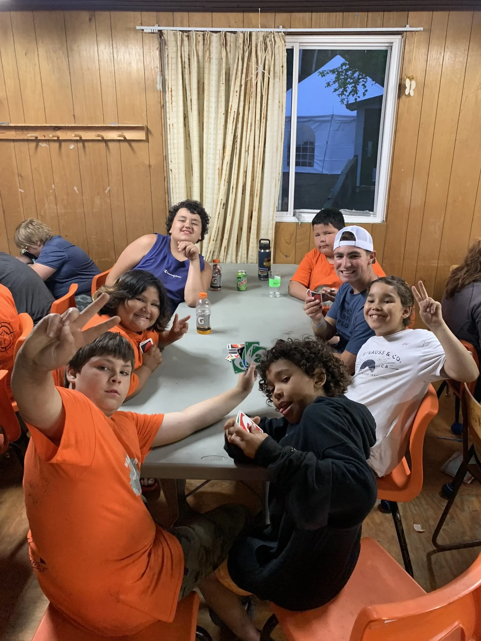 Group of people playing a card game at a table indoors, with some making peace signs and showing friendly gestures.
