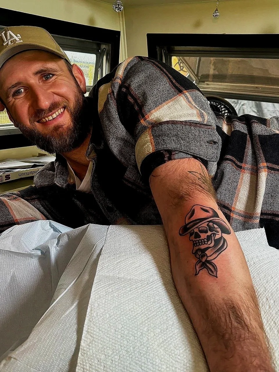 Un homme souriant montrant son bras tatoué d'un squelette cowboy avec un chapeau et un bandana, dans la caravan.