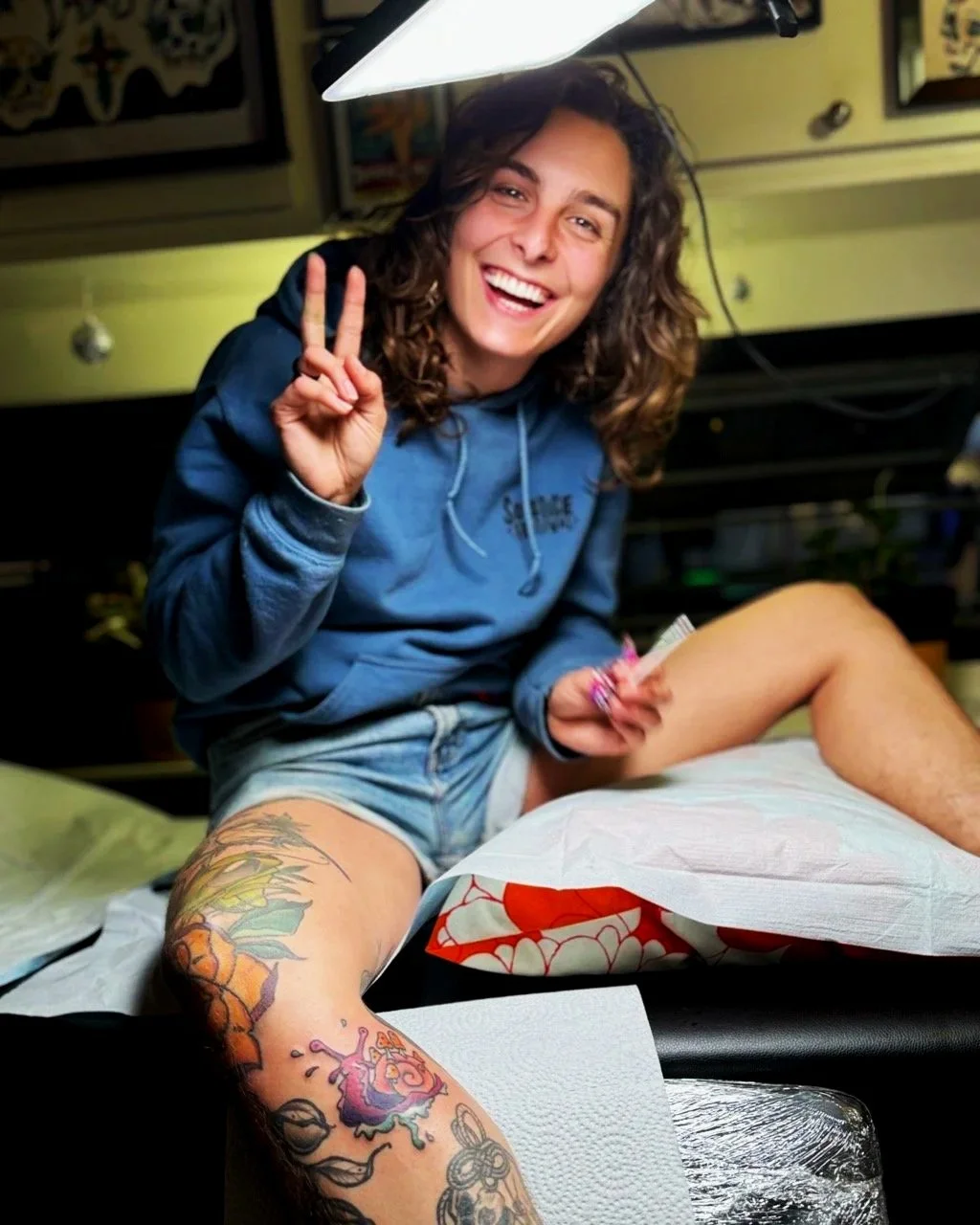 Une femme souriante faisant un signe de paix avec la main, assise avec une jambe tatouée, dans un environnement intérieur lumineux.