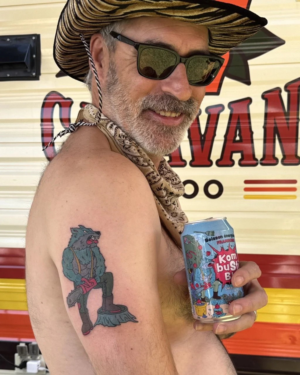 Homme souriant portant un chapeau tressé, lunettes de soleil, avec un tatouage d'un loup en costume de cowboy sur le bras, tenant une canette de boisson énergisante colorée avec un dessin de loup dans un style cartoon. 