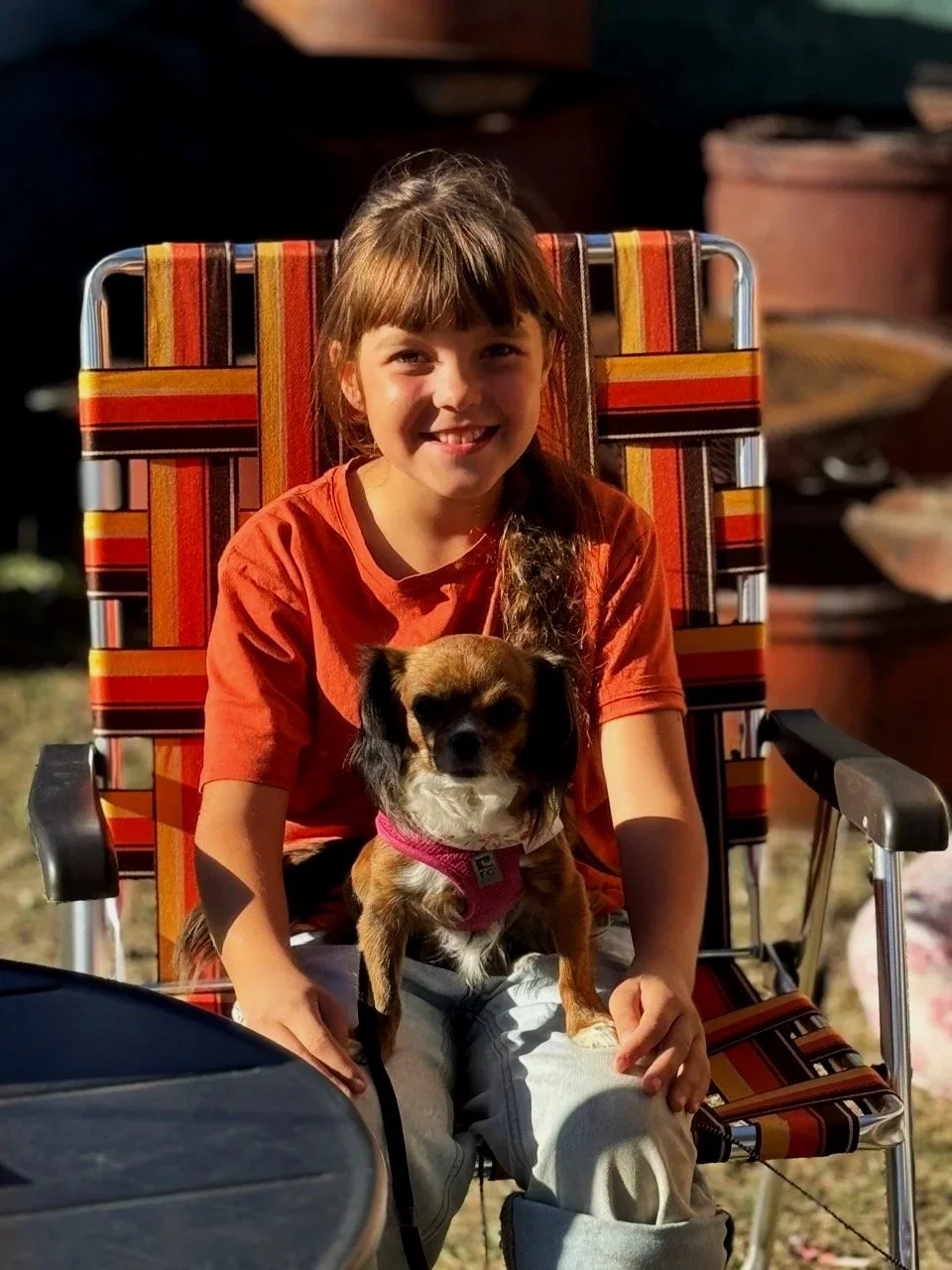 une fille rousse assise sur une chaise en extérieur, tenant un petit chien brun avec des oreilles noires, tous deux regardant l'appareil photo, dans un cadre en plein air avec des éléments de mobilier