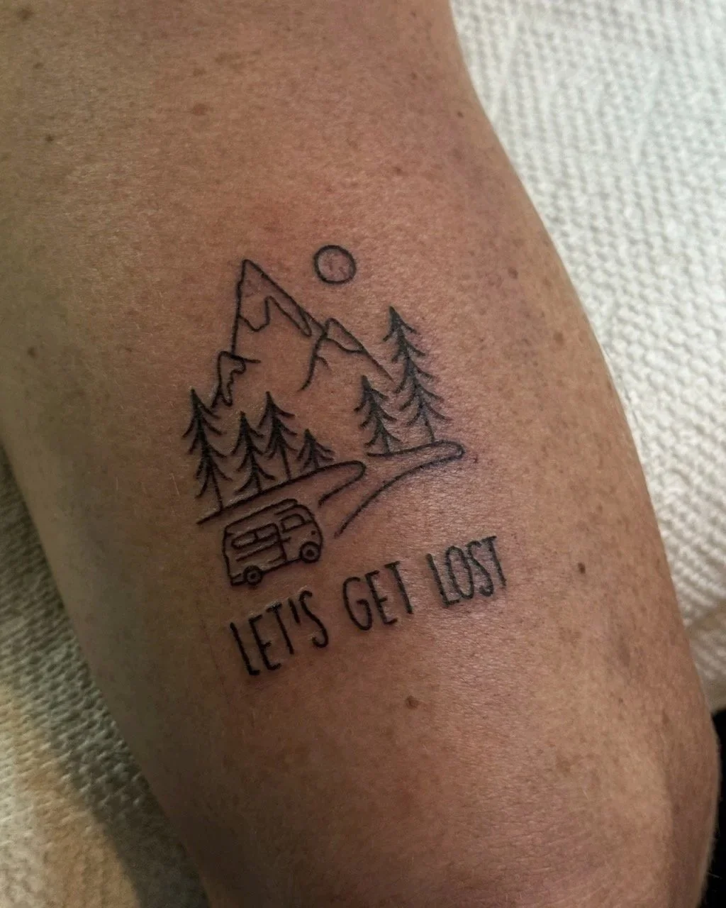 Tatouage représentant une montagne, des arbres, une route et une voiture avec le texte 'Let's get lost'.