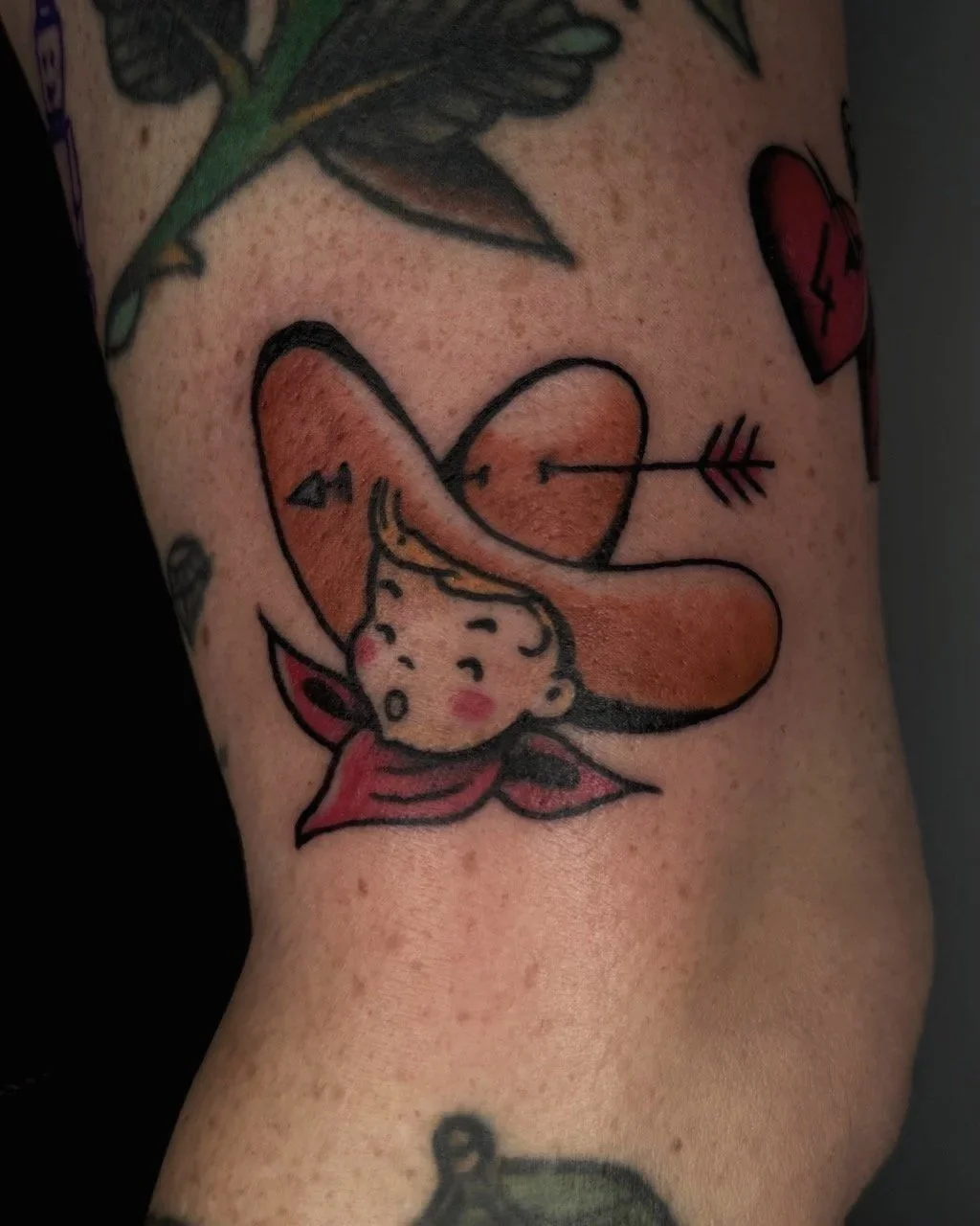 Tatuage d'une petite gars avec un large chapeau orange et un bandana rouge, un cœur avec une flèche, sur la peau.
