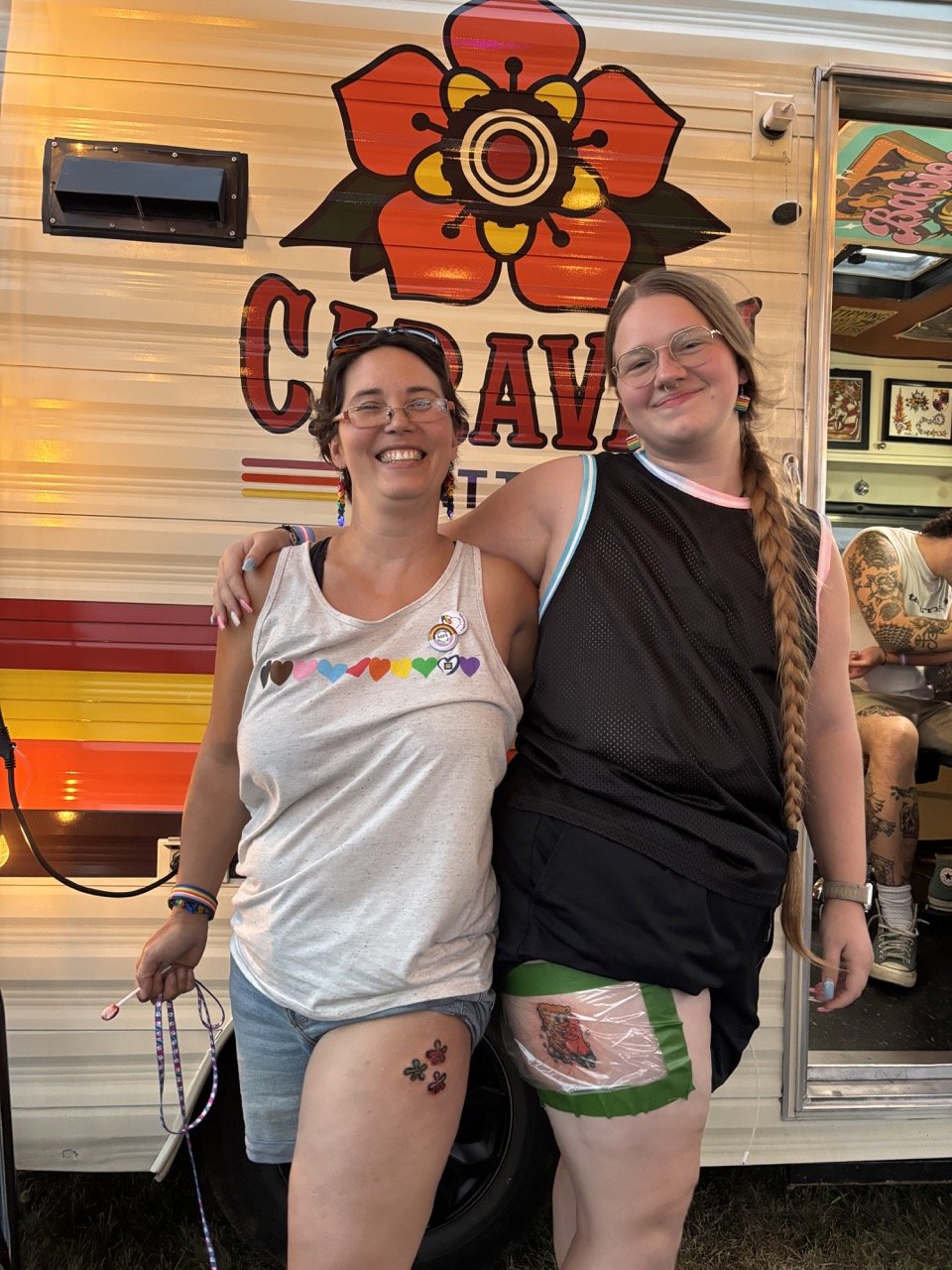 Deux femmes souriantes, debout côte à côte, posant devant la caravan coloré. Elles portent des vêtements décontractés, avec des tattoos visibles sur leurs jambes et des accessoires colorés.