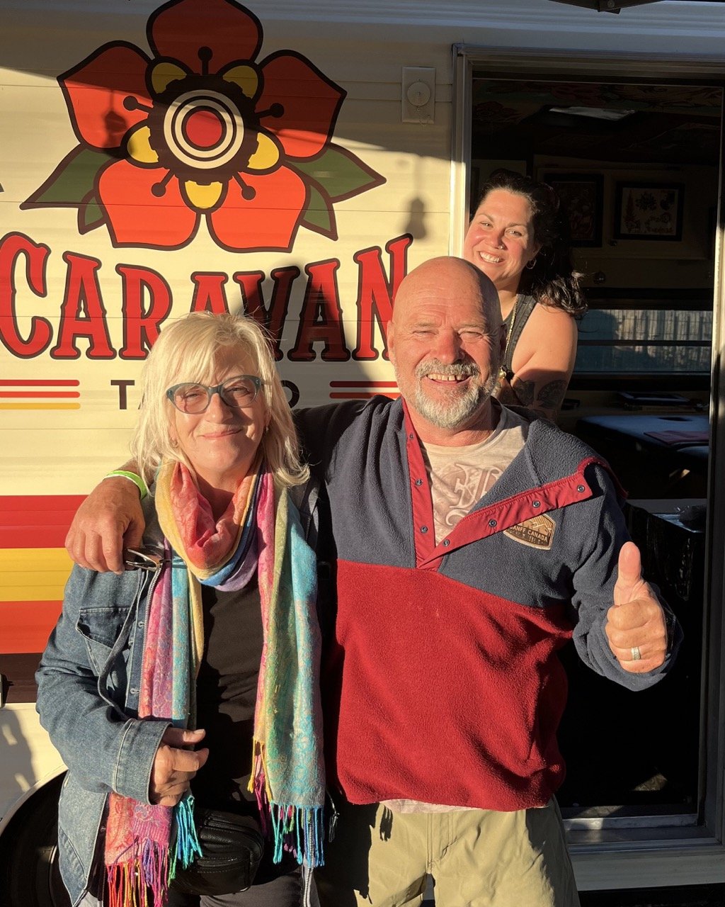 Trois personnes souriantes posent devant un food truck nommé "CARAJAN", avec un grand logo floral au-dessus du nom, en plein jour.