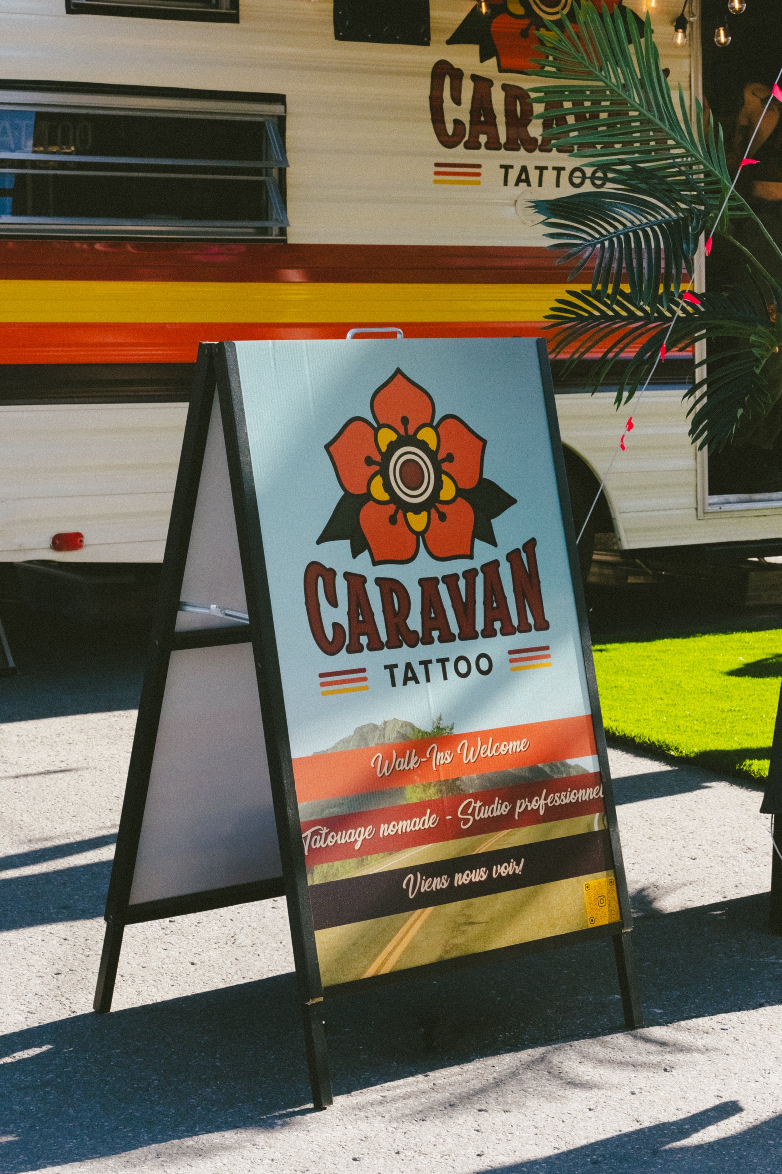 Panneau indiquant l'emplacement d'un tatoueur nommée Caravan Tattoo, avec une image de fleur et un fond de paysage désertique, invitant les passants à entrer, dans un environnement en plein air avec une caravane en arrière-plan.