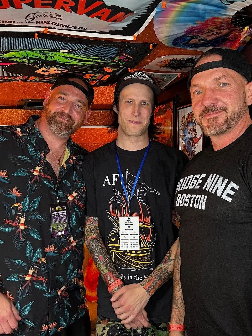 Trois hommes posent pour une photo, ils portent des casquettes ou chapeaux et des vêtements avec des motifs. Le décor est coloré, avec des posters ou des affiches derrière eux.