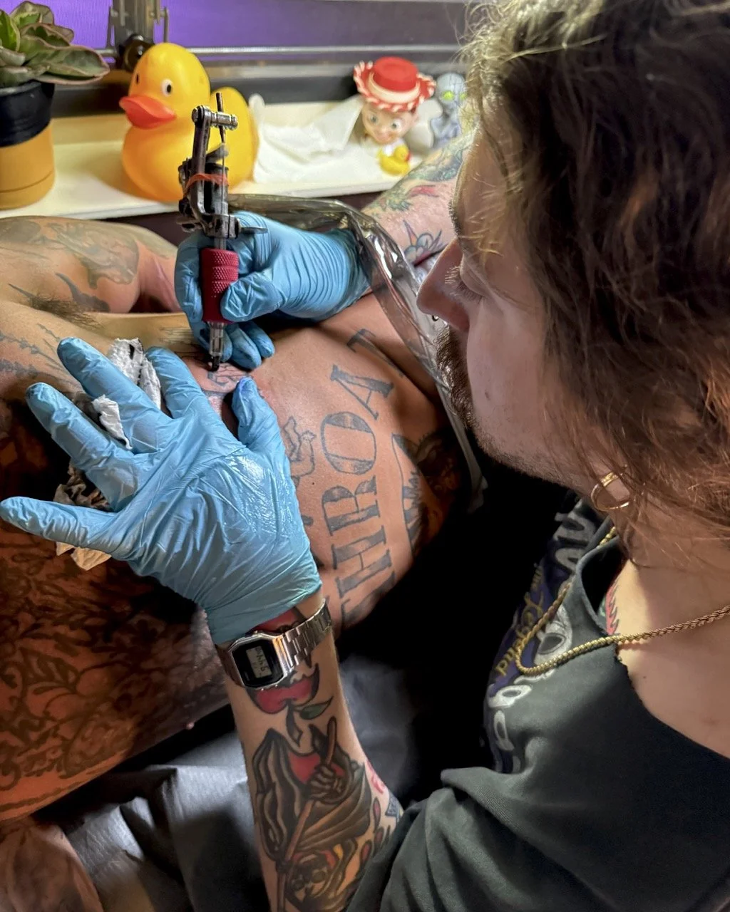 Un tatoueur en train de tatouer un bras, portant des gants bleus, avec des objets de décoration en arrière-plan, y compris un canard en plastique jaune et une figurine d'enfant.