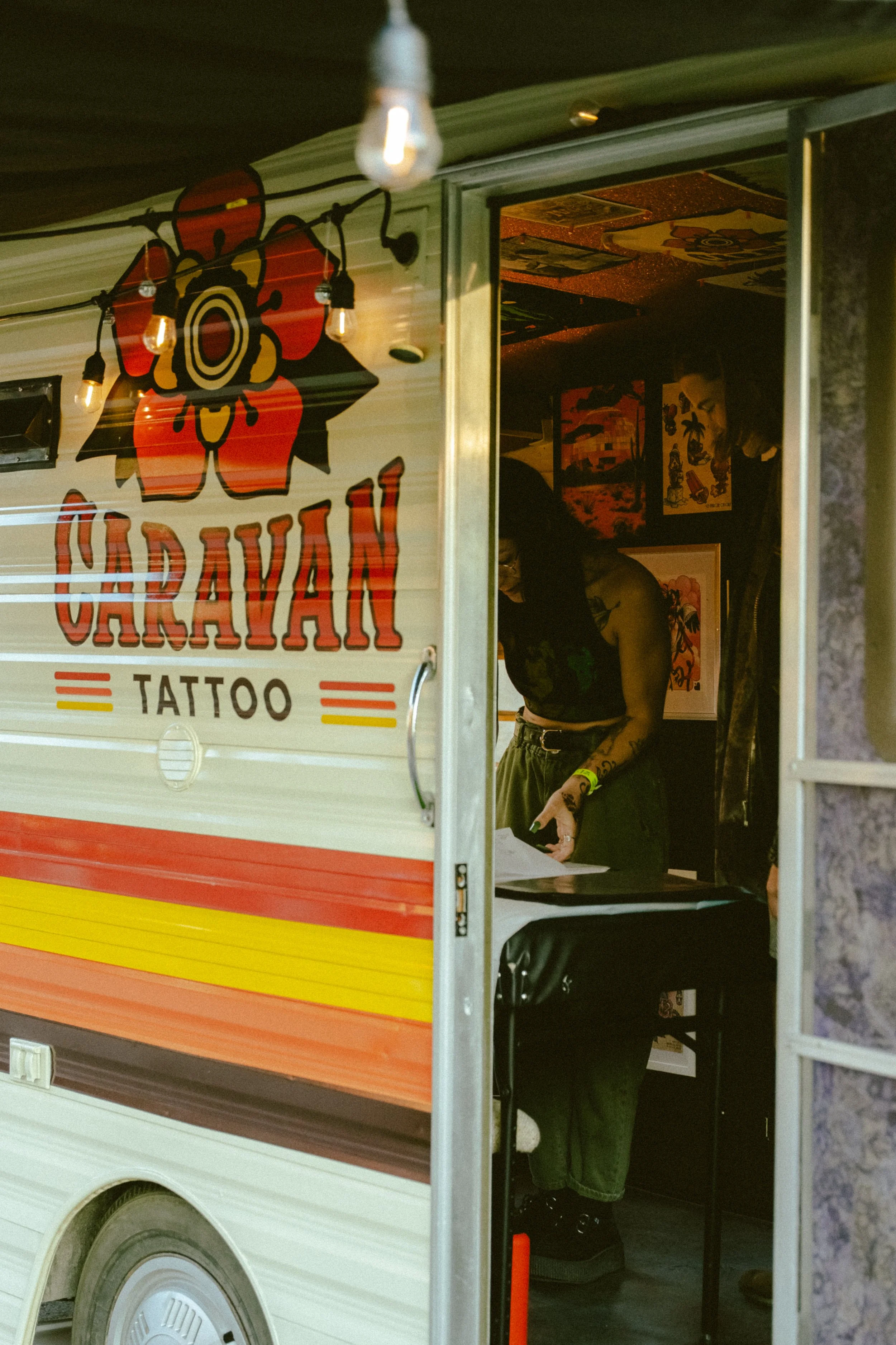 Image de la caravan avec le gros logo de l'entreprise.  La porte ouverte est ouverte et on y voit une tatoueuse