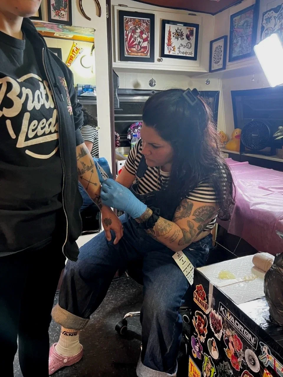 Une femme est en train de se faire tatouer le bras par une autre personne dans une pièce décorée de dessins et affiches colorés.