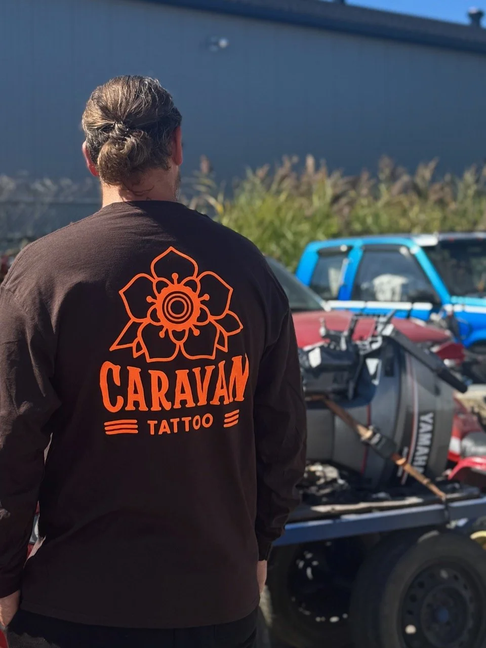 Un homme vu de dos portant un blouson noir avec un logo orange et le texte « CARAVAN TATTOO » sur le dos, derrière un véhicule en remorque.