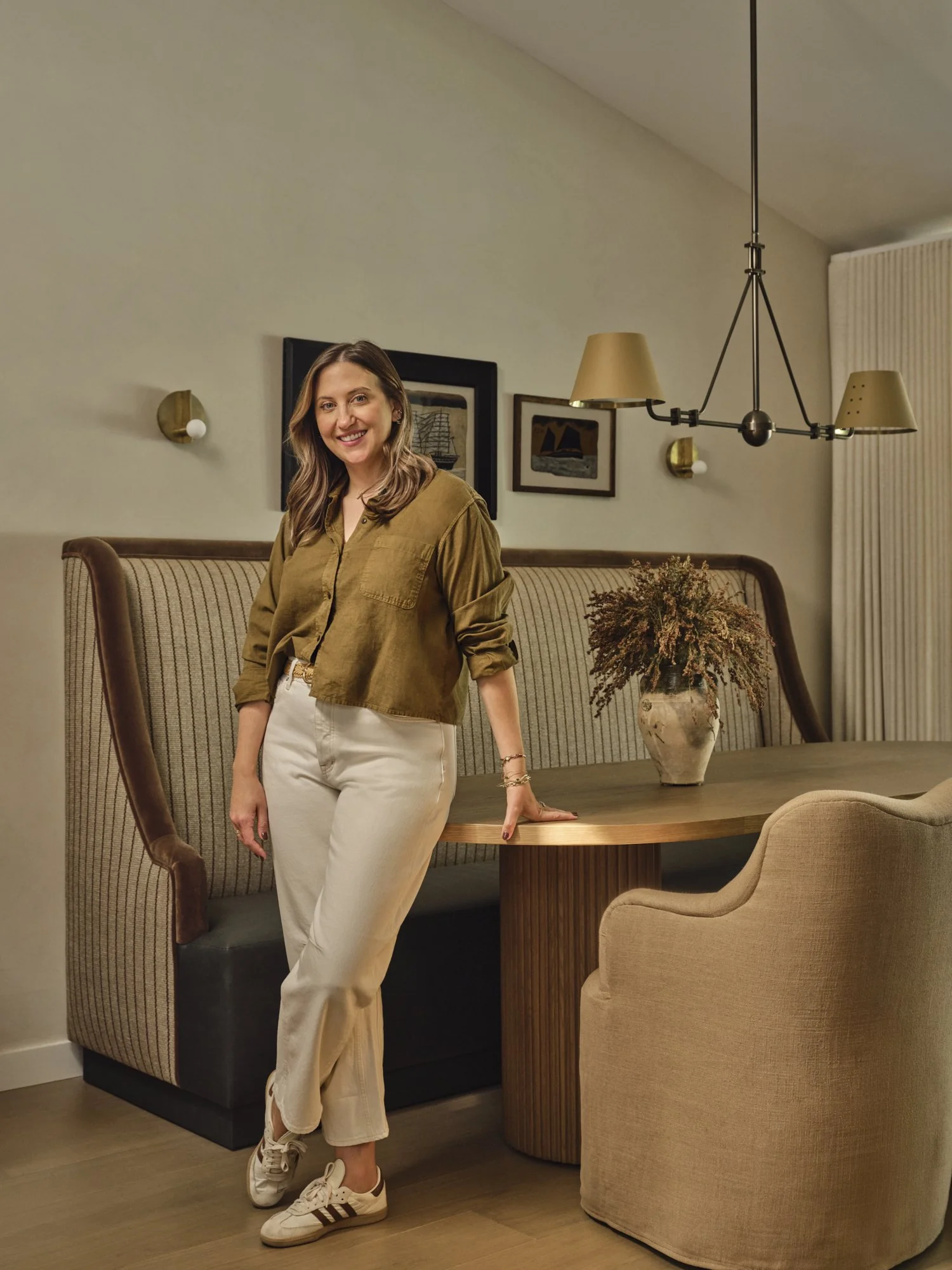 A woman with long blonde hair wearing a brown jacket, white pants, and Adidas sneakers standing in a dining room with a table, vases, and framed artwork on the wall.