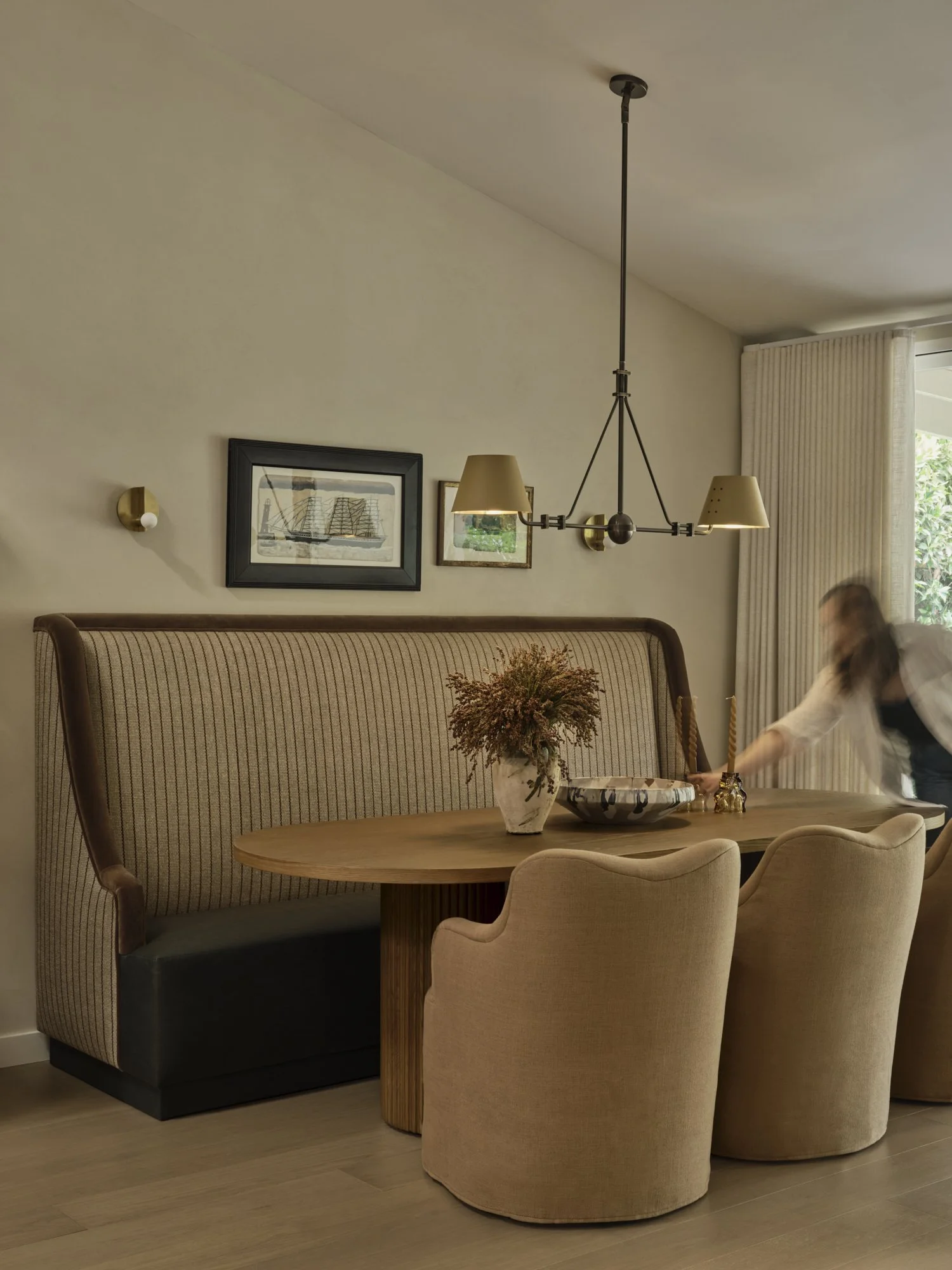 Dining room with a wooden table, beige upholstered chairs, a vase with dried flowers, two framed pictures of ships on the wall, gold wall sconces, and a modern chandelier. There are curtains over a window to the right, and a person is reaching on the