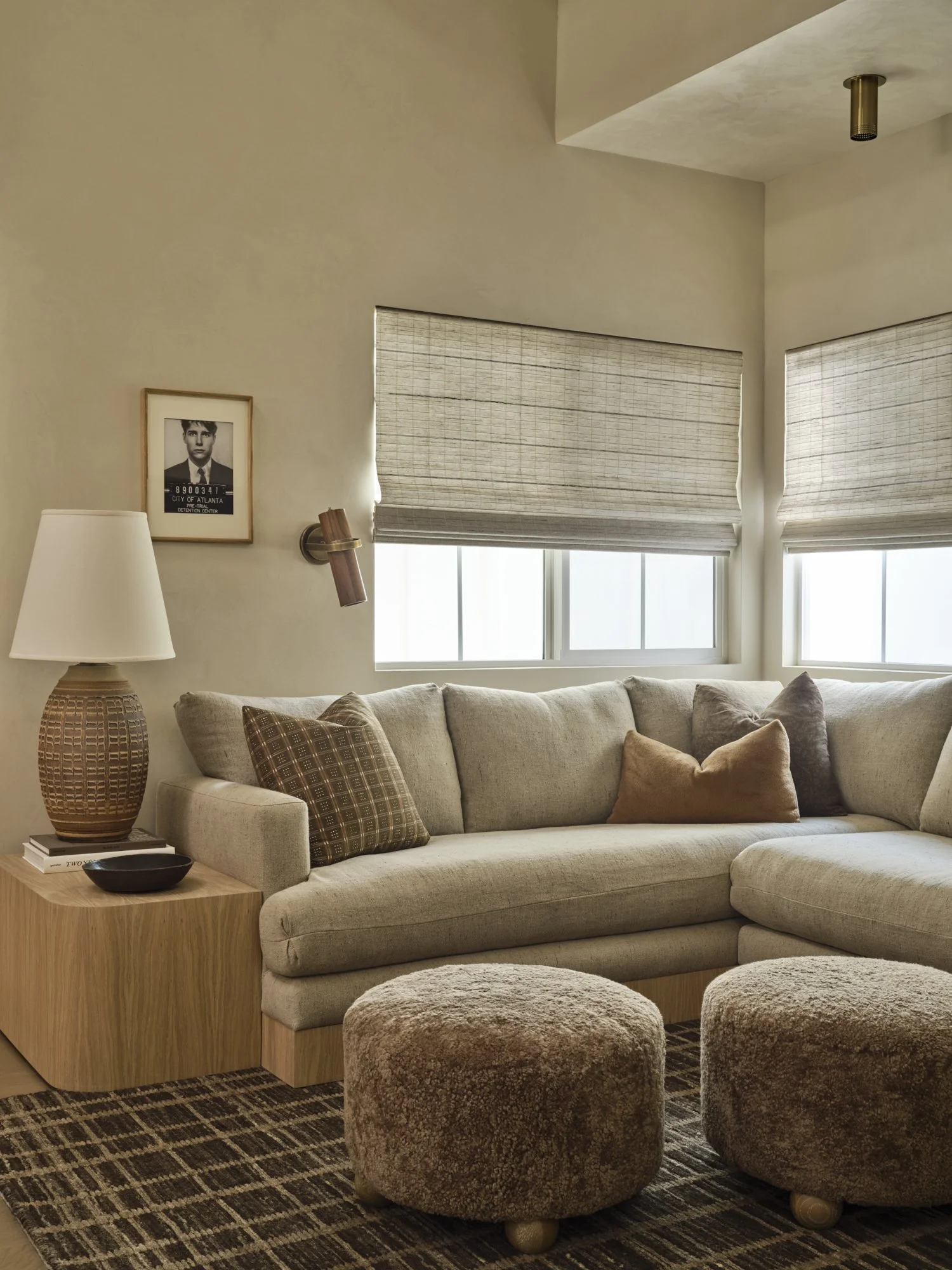 Living room with beige sofa, decorative pillows, wooden side table with lamp, picture frame on wall, bean bag ottomans, curtains, patterned rug.