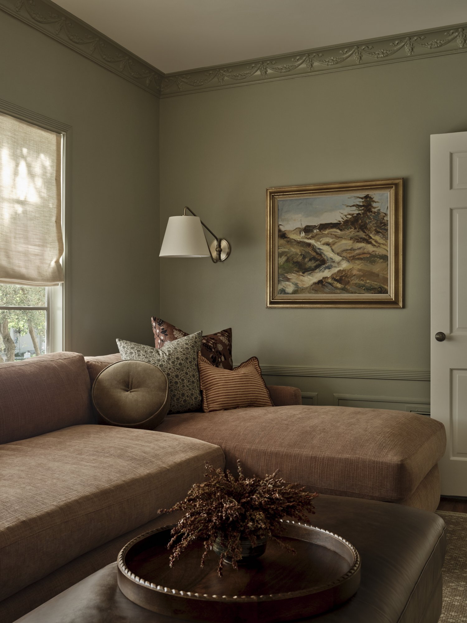 A cozy living room corner with a pink sectional sofa, decorative pillows, a floor lamp, framed landscape painting, and a tray table with dried flowers, near a window with beige blinds.