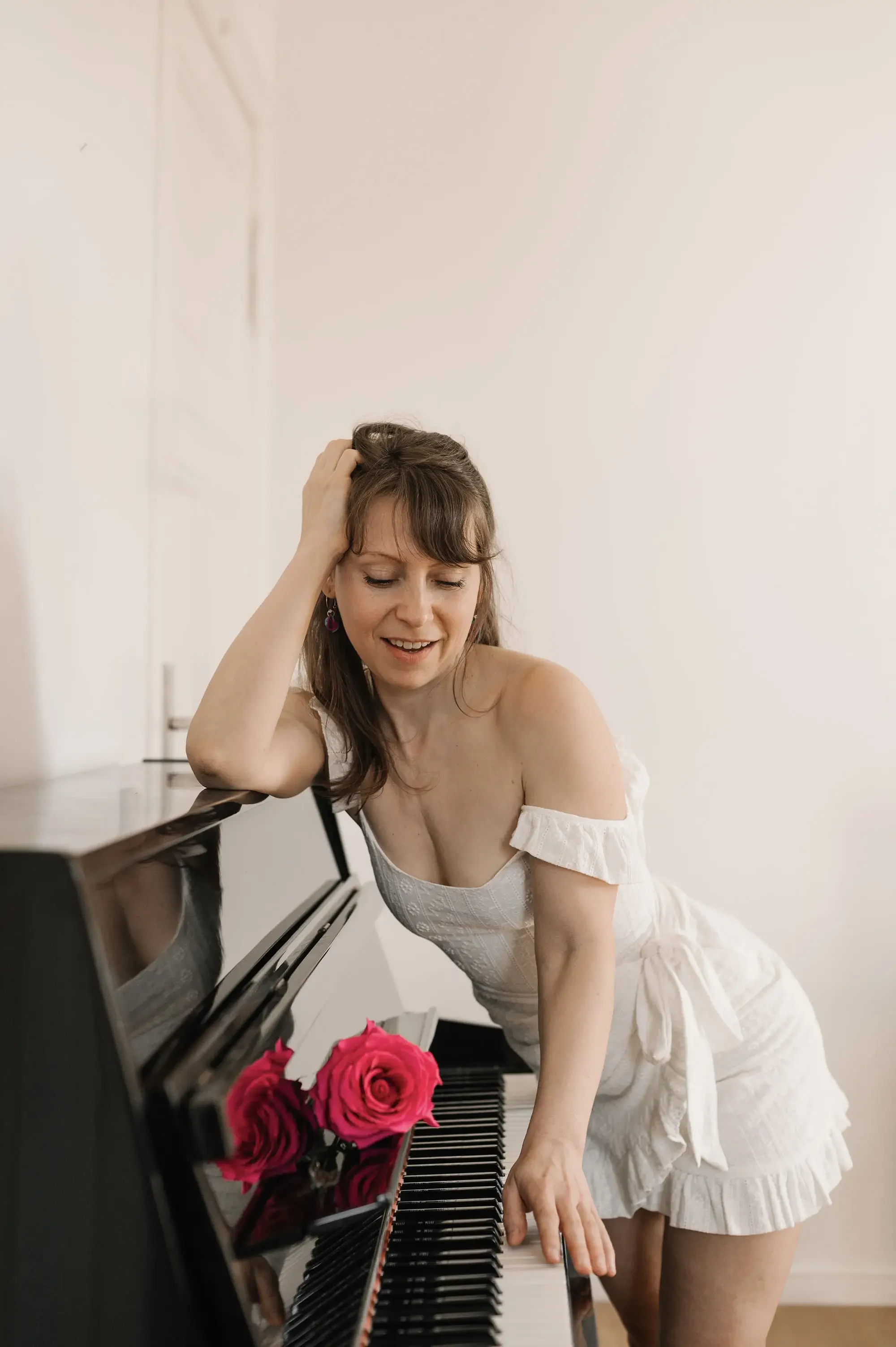 Junge Frau in Weißes Kleid, die an einem schwarzen Klavier sitzt, mit pinken Rosen auf dem Klavier, lächelnd und die Stirn berührend.