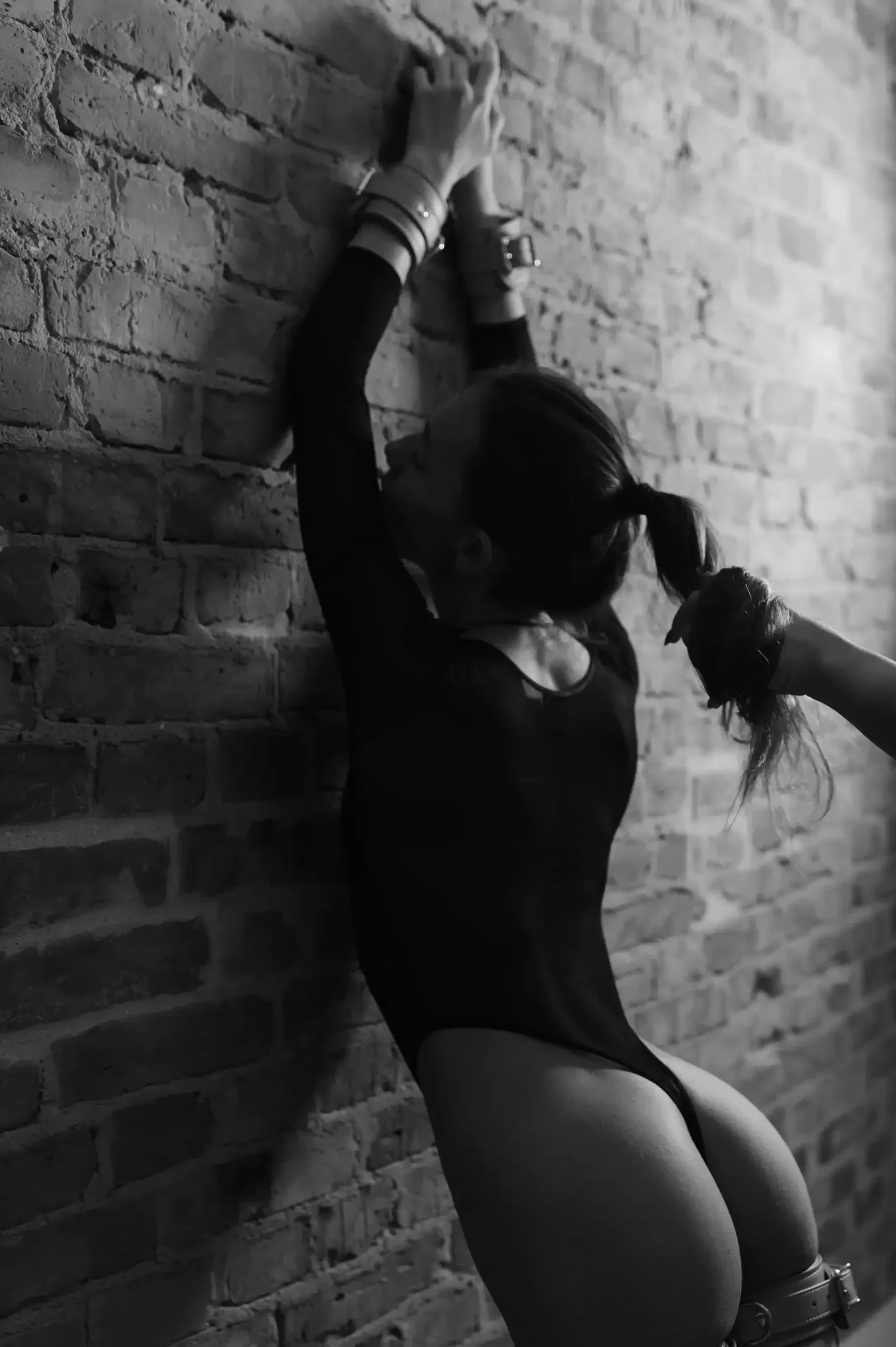 A woman in a black outfit with long sleeves and BDSM-style cuffs on her wrists is standing against a brick wall. She is holding a lock of her own hair with her right hand and appears to be licking or kissing the wall. The photo is in black and white.