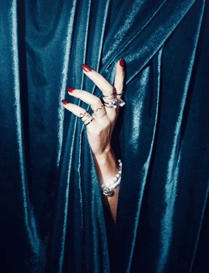 Hand mit roten Nägeln und mehreren Ringen, die durch einen Spalt in einem dunkelblauen, schimmernden Vorhang greift.