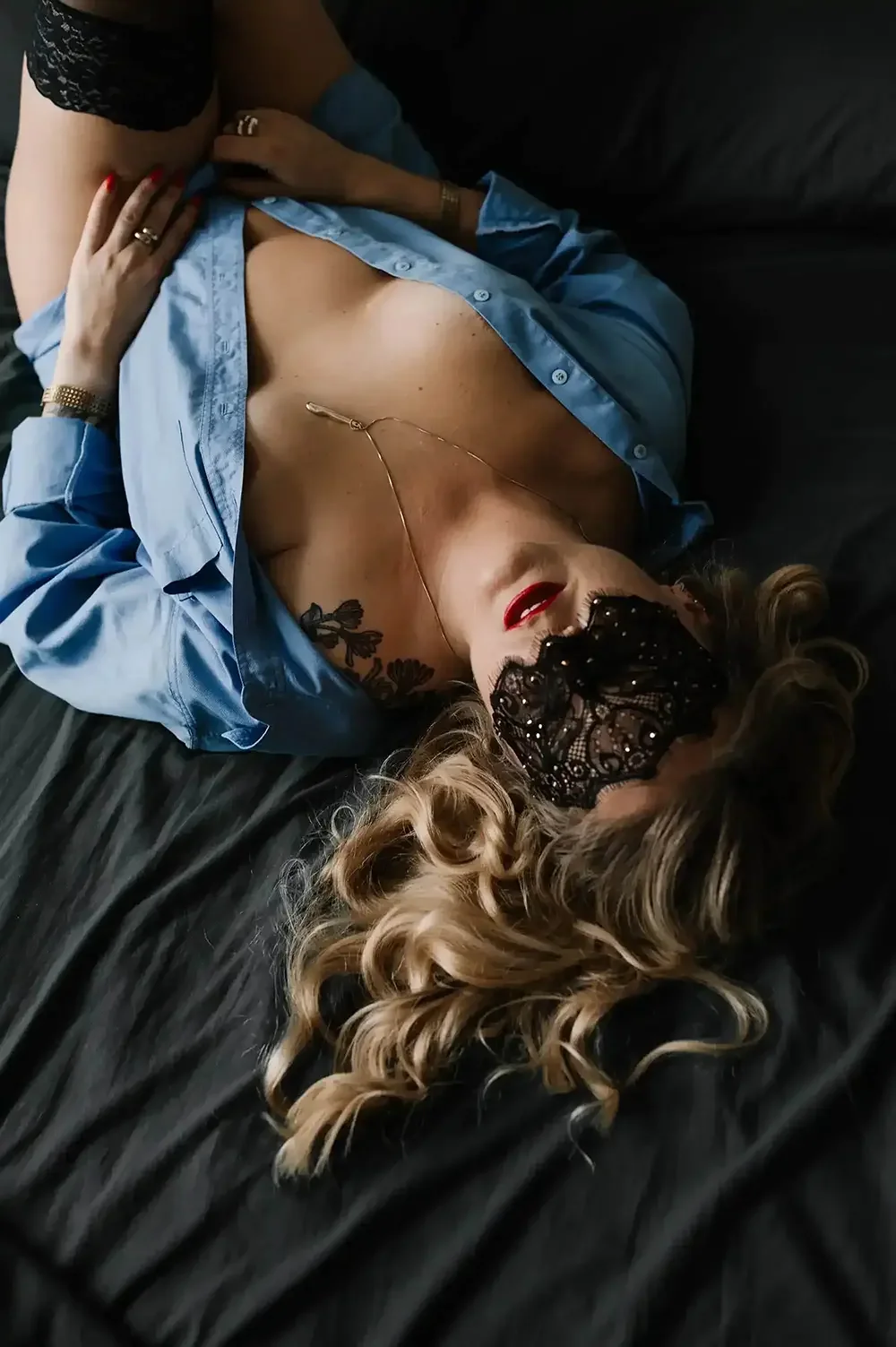 Frau mit blonden Locken, schwarzer Spitze-Maske, roten Lippen, blauer Seidenbluse, liegt auf schwarzem Bett, trägt eine Halskette und ein Tattoo auf der Brust.