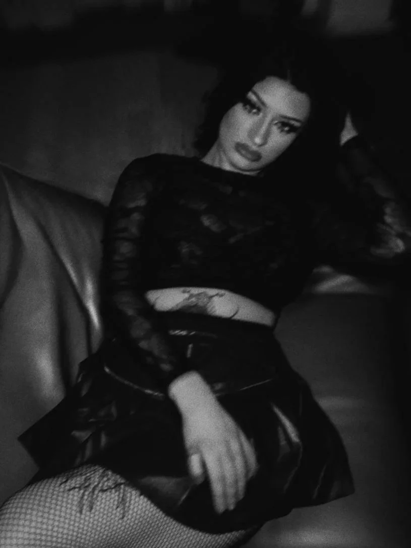 Schwarz-weiße Fotografie einer jungen Frau mit dunklem, lockigem Haar, die auf einem Sofa liegt, schwarzes Oberteil mit transparenten Details trägt, und an der Kamera vorbei schaut.