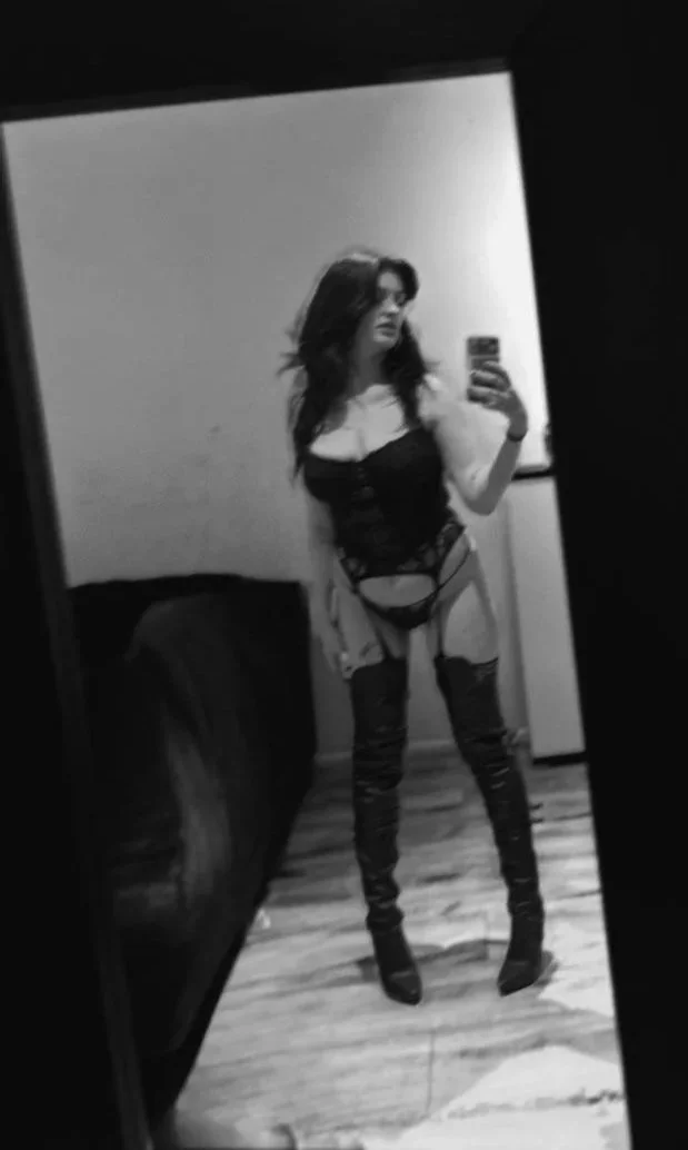 Eine Frau nimmt ein Selfie in einem Raum auf einem Spiegel auf. Sie trägt schwarze Lingerie und Overknee-Stiefel.