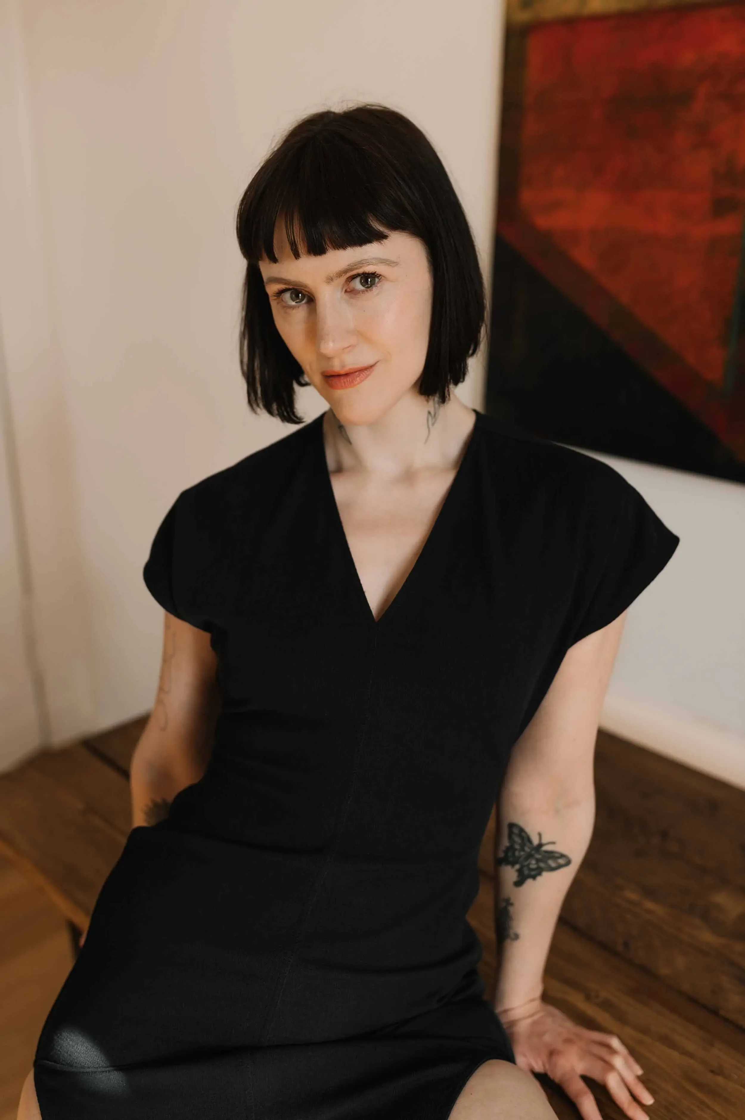 Junge Frau mit schwarzem Bob-Haarschnitt, schwarzen Kleid, Tattoos am Arm, sitzt auf einem Tisch vor weißer Wand und bemaltem Kunstwerk.