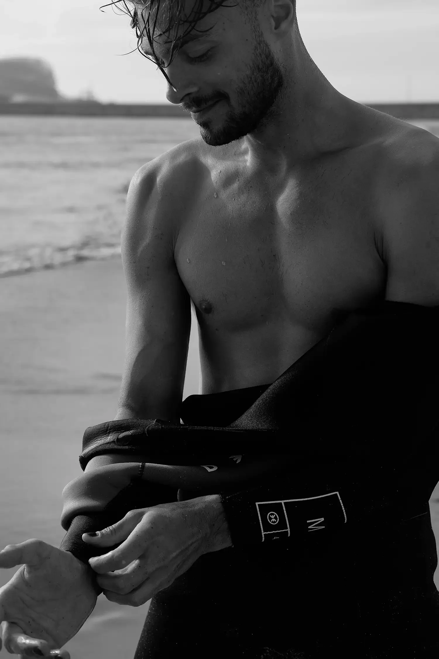 Ein nackter Mann steht am Strand mit Wasser im Hintergrund, während er seine Jacke anzieht.