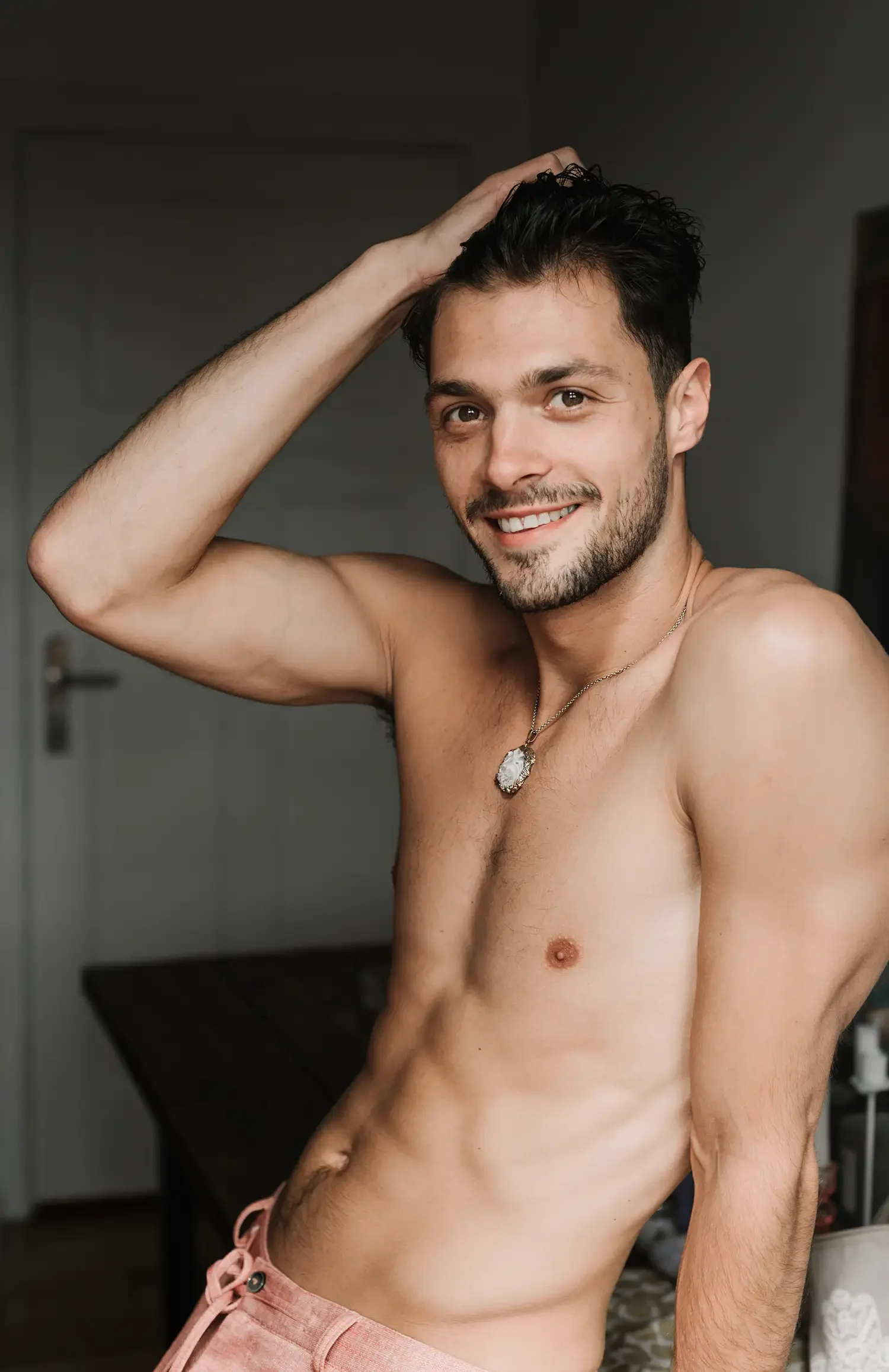 Ein lachender, nackter junger Mann mit dunklem, lockigem Haar, kurzen Bart, trägt eine Halskette mit Zahnmedaillon, steht in einem Raum mit dunklem Hintergrund.