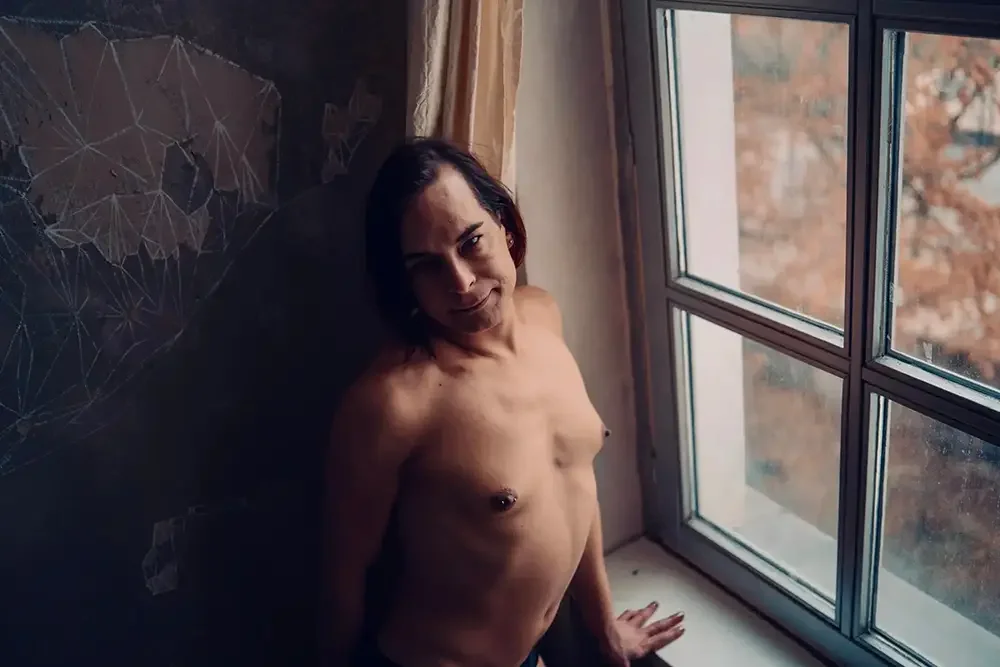 Eine nackte Frau mit dunklen Haaren sitzt auf dem Boden neben einem Fenster, das eine winterliche Herbstlandschaft zeigt.