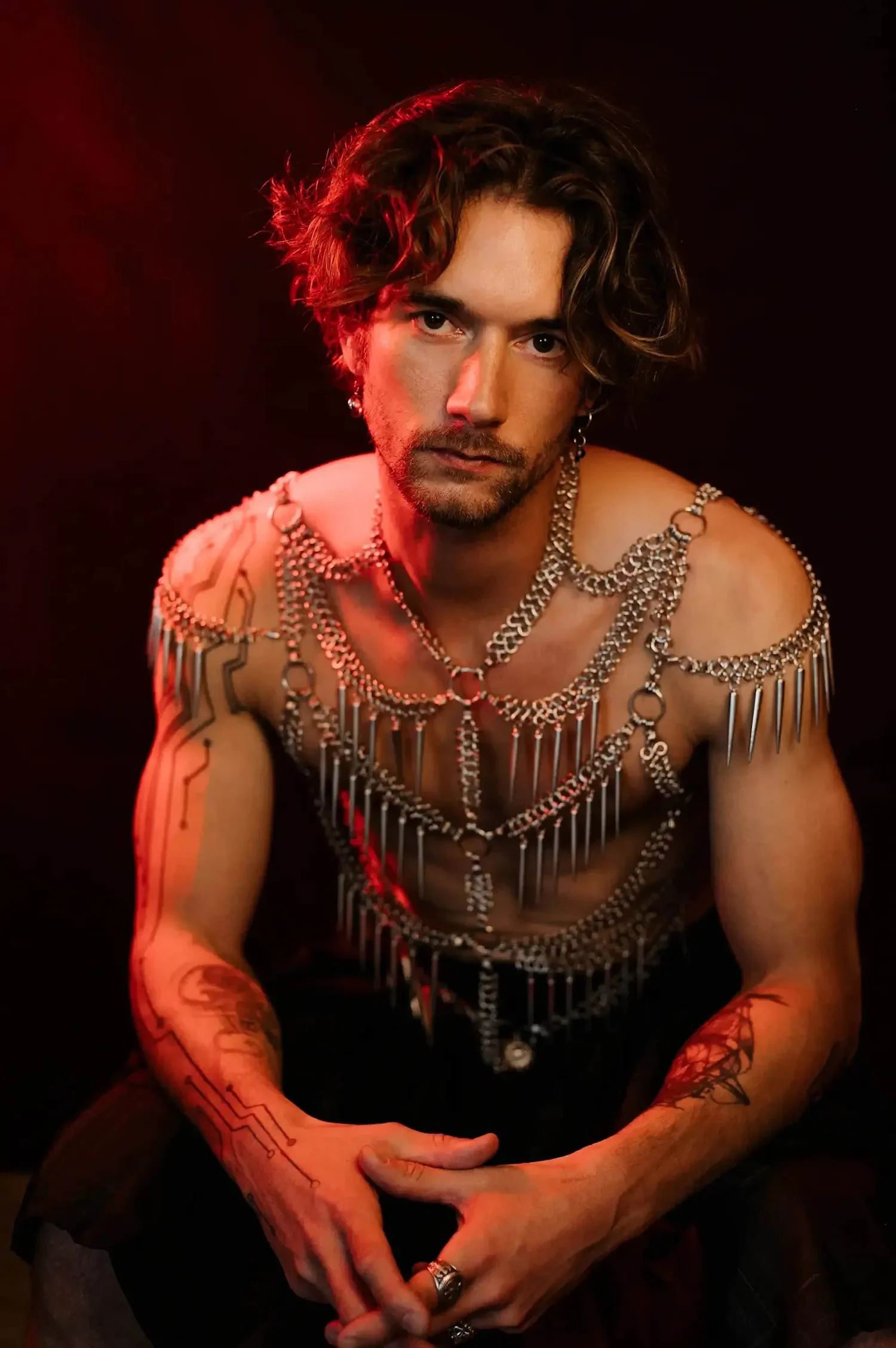 Junger Mann mit Tätowierungen und Schmuck, trägt mehrere Ketten, sitzt vor einem dunklen Hintergrund, mit rotem Lichtschein